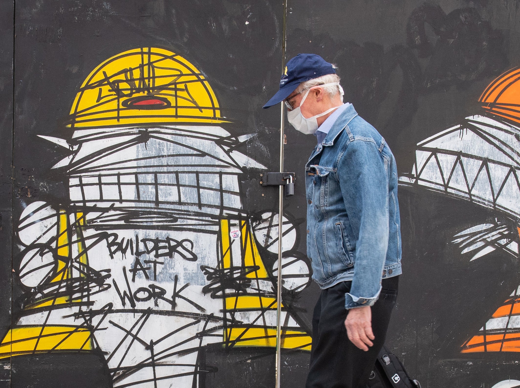 A man wearing a protective face mask walks past a street art mural showing construction workers in central London