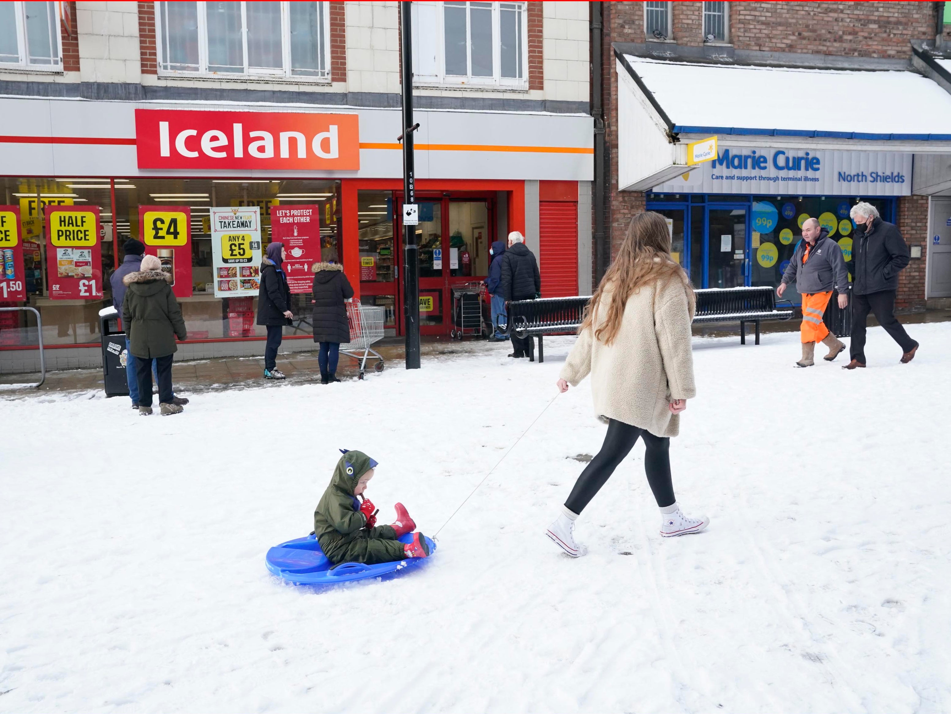 A woman pulls a child on a sledge past a line of people waiting in the snow outside and Iceland store in North Shields, Tyne and Wear, as the cold snap continues to grip much of the nation. Picture date: Friday February 12, 2021.