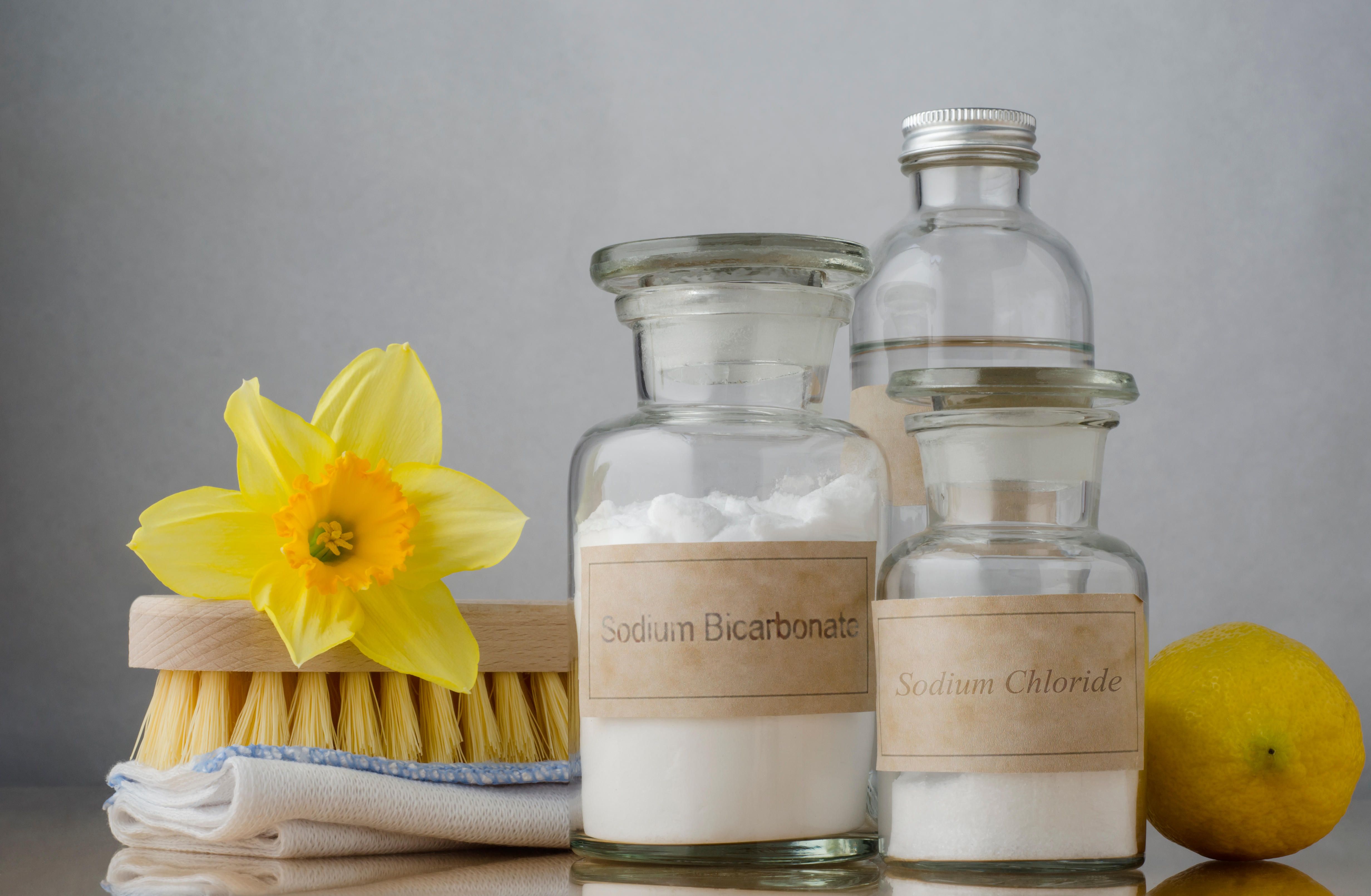 Still life of natural cleaning choices. Sodium bicarbonate and salt in apothecary jars, white vinegar behind them and a lemon to the right. A folded