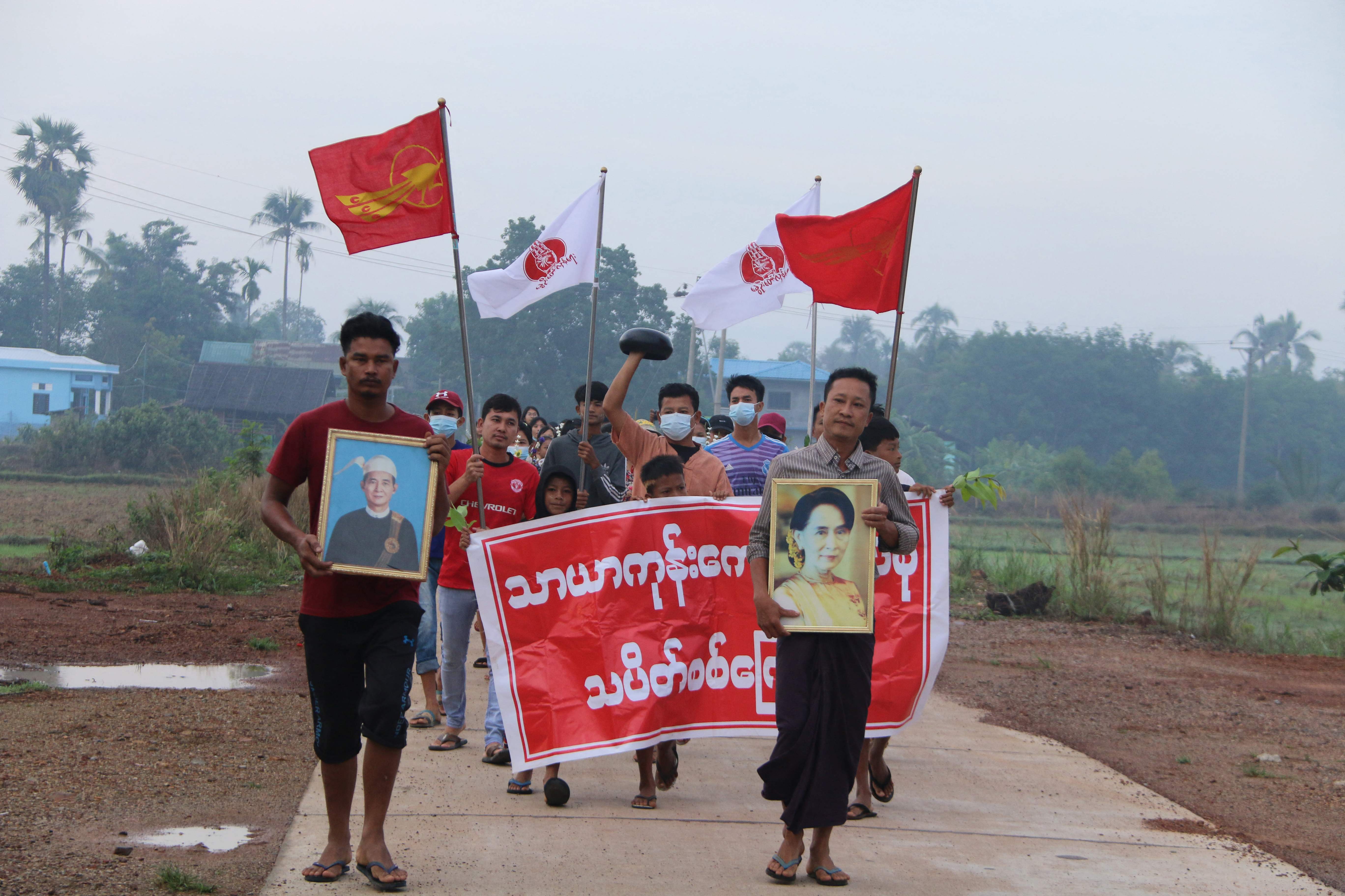 This handout photo taken and released by Dawei Watch on April 14, 2021 shows protesters carrying flags and a portrait of detained Myanmar civilian leader Aung San Suu Kyi (R) as they march during a demonstration against the military coup in Dawei