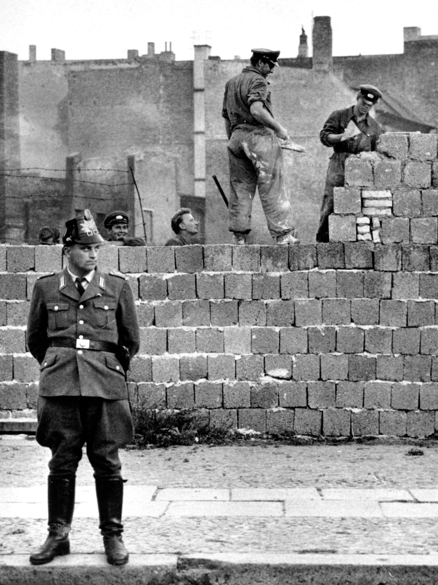 October 1961: a West Berlin police officer stands in front of a concrete wall dividing East and West Berlin at Bernauer Strasse as East Berlin workers add blocks to the wall to make it higher