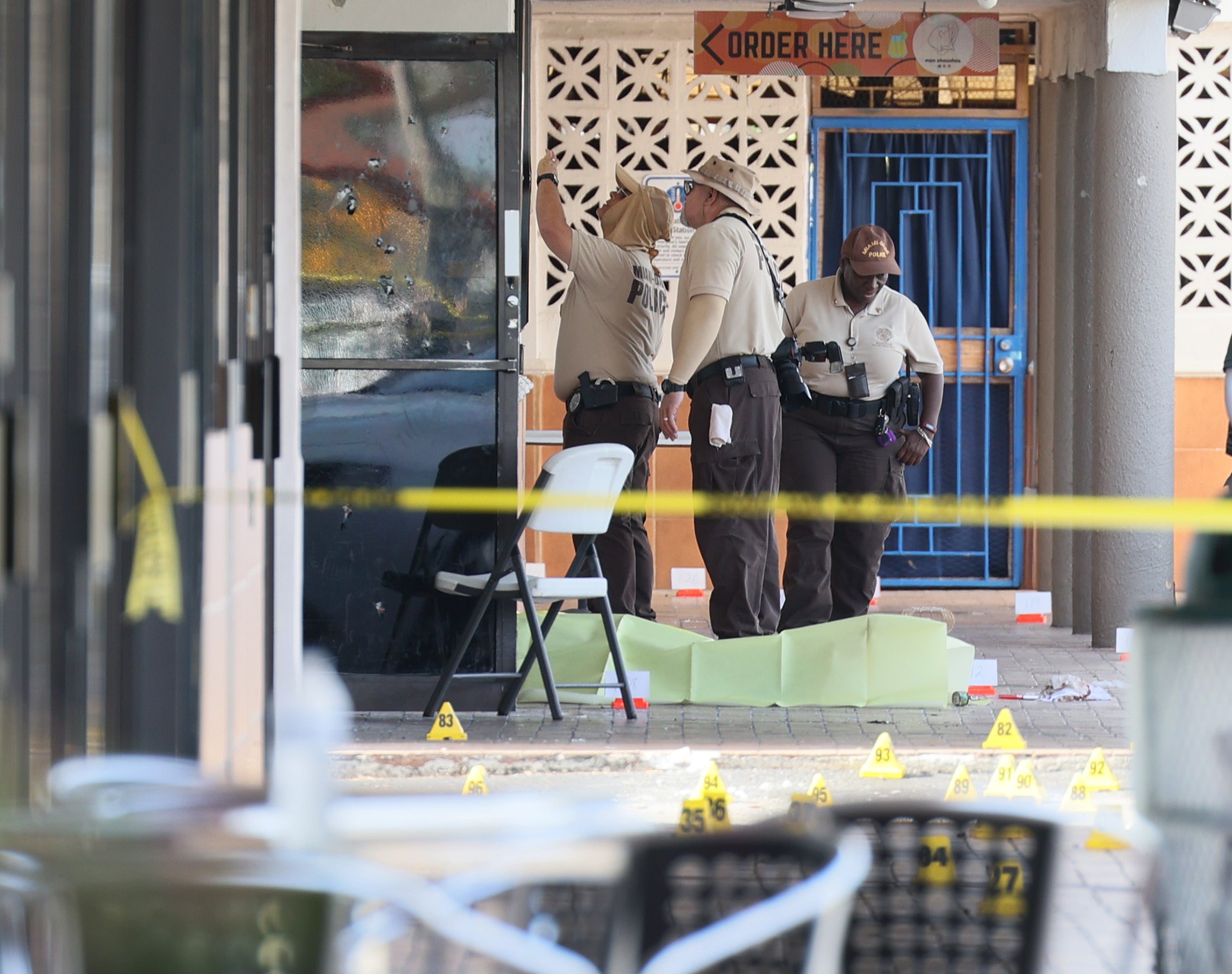 Miami-Dade police investigate near shell case evidence markers on the ground and a door with what appear to be bullet holes