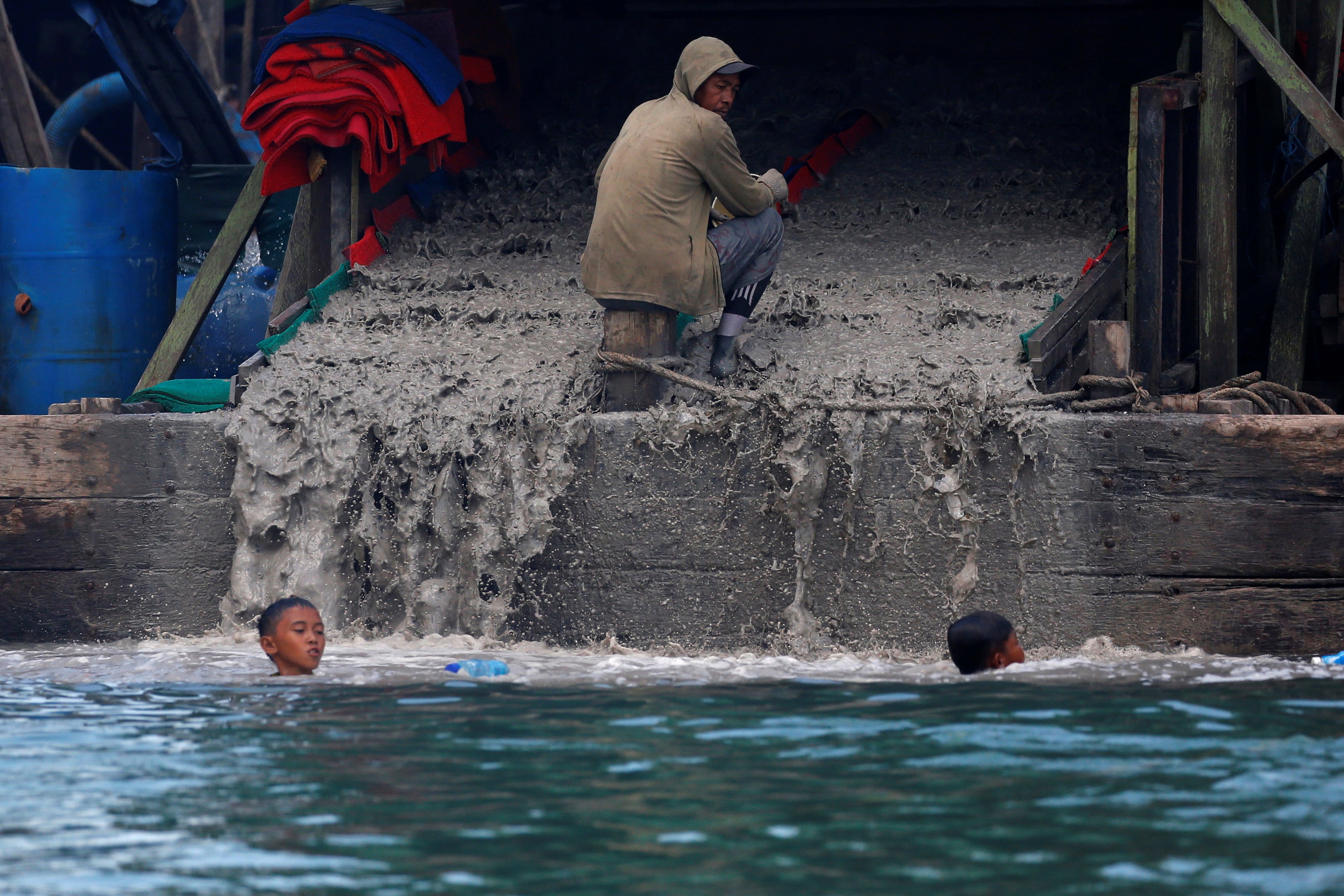 An artisanal tin miner rests as children swim near a pontoon