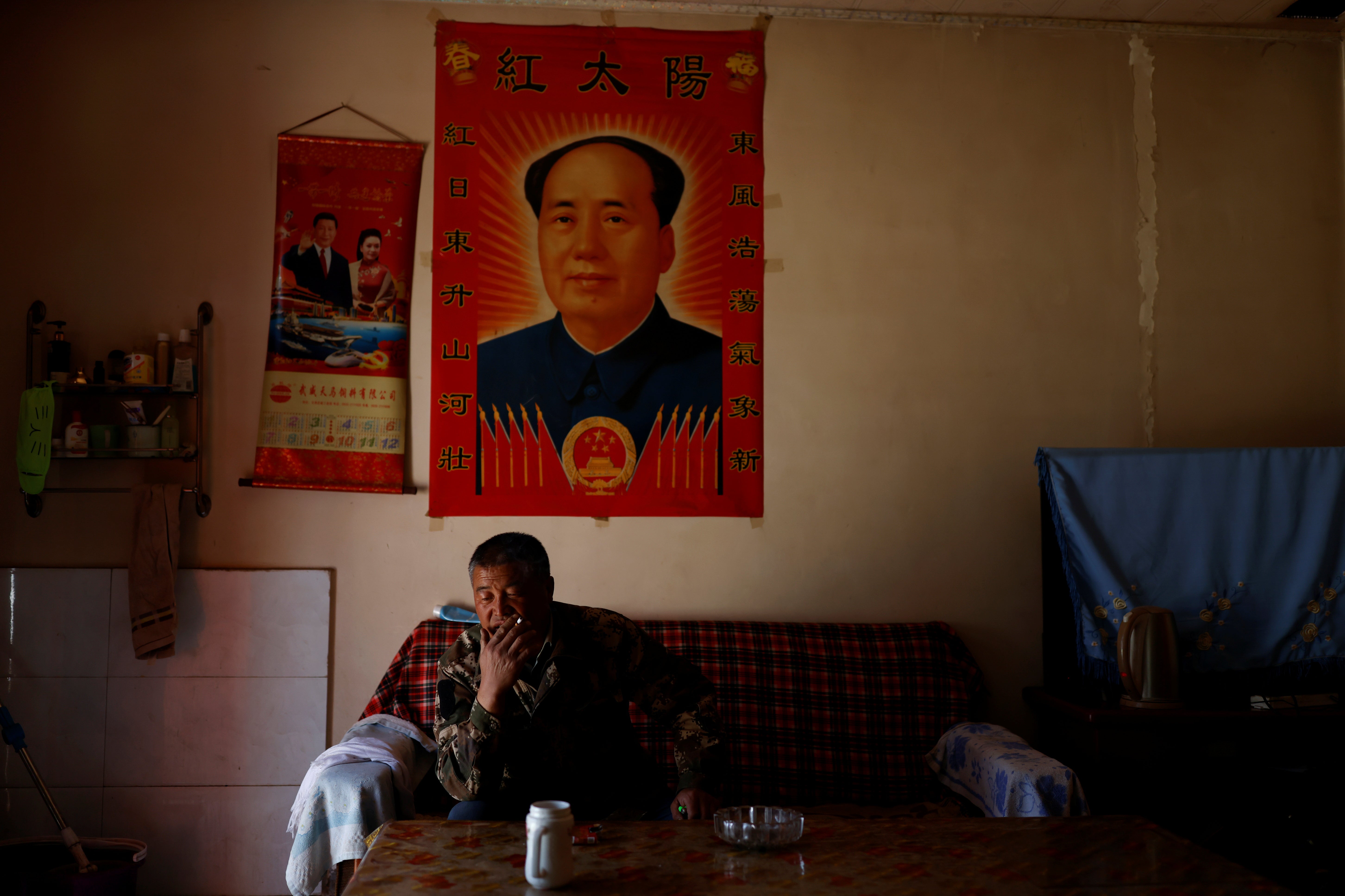 Wang Yinji sits in front of posters of late Chinese chairman Mao Zedong and president Xi Jinping and his wife Peng Liyuan as he smokes at his house