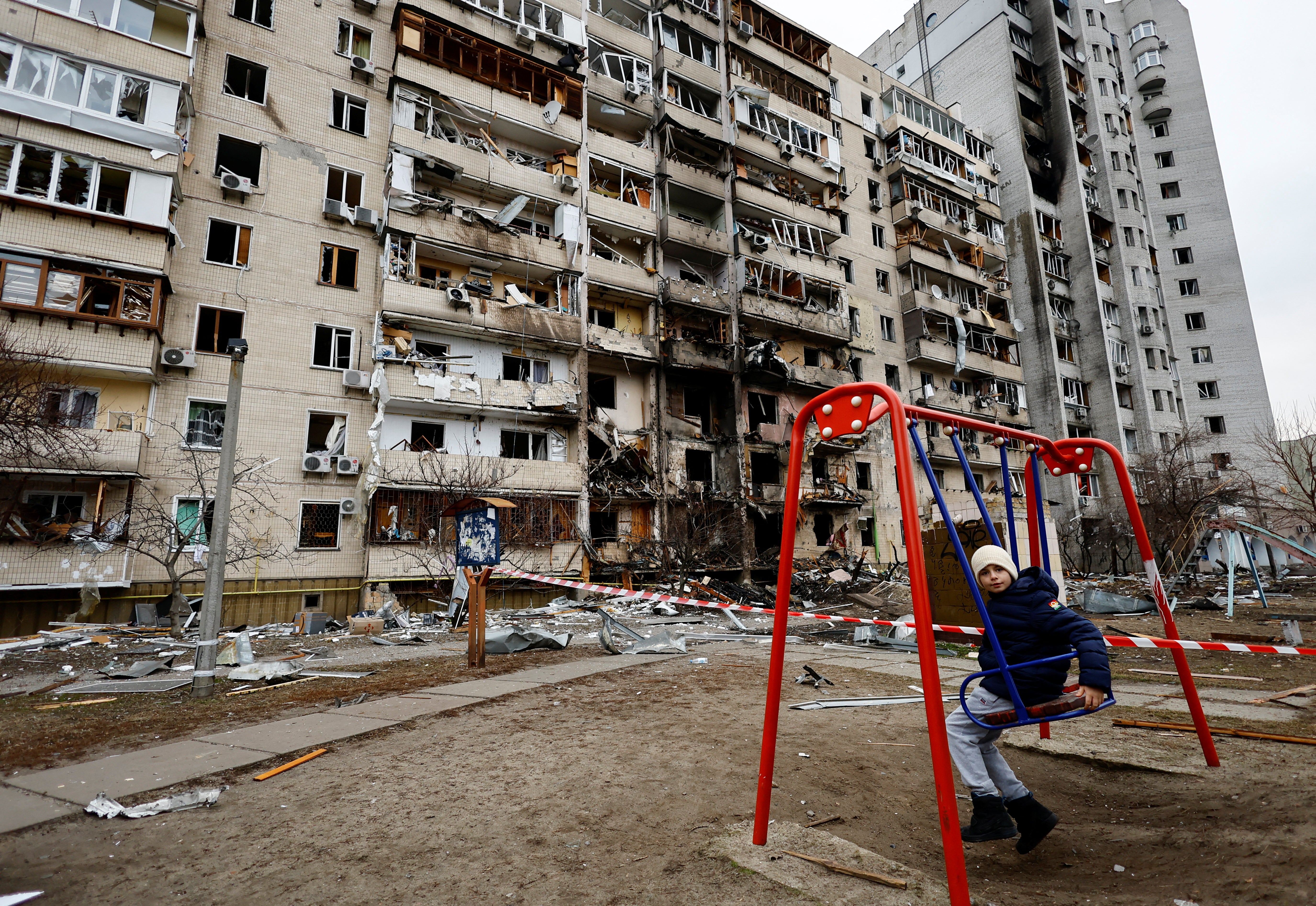 A child sits on a swing in front of a damaged residential building