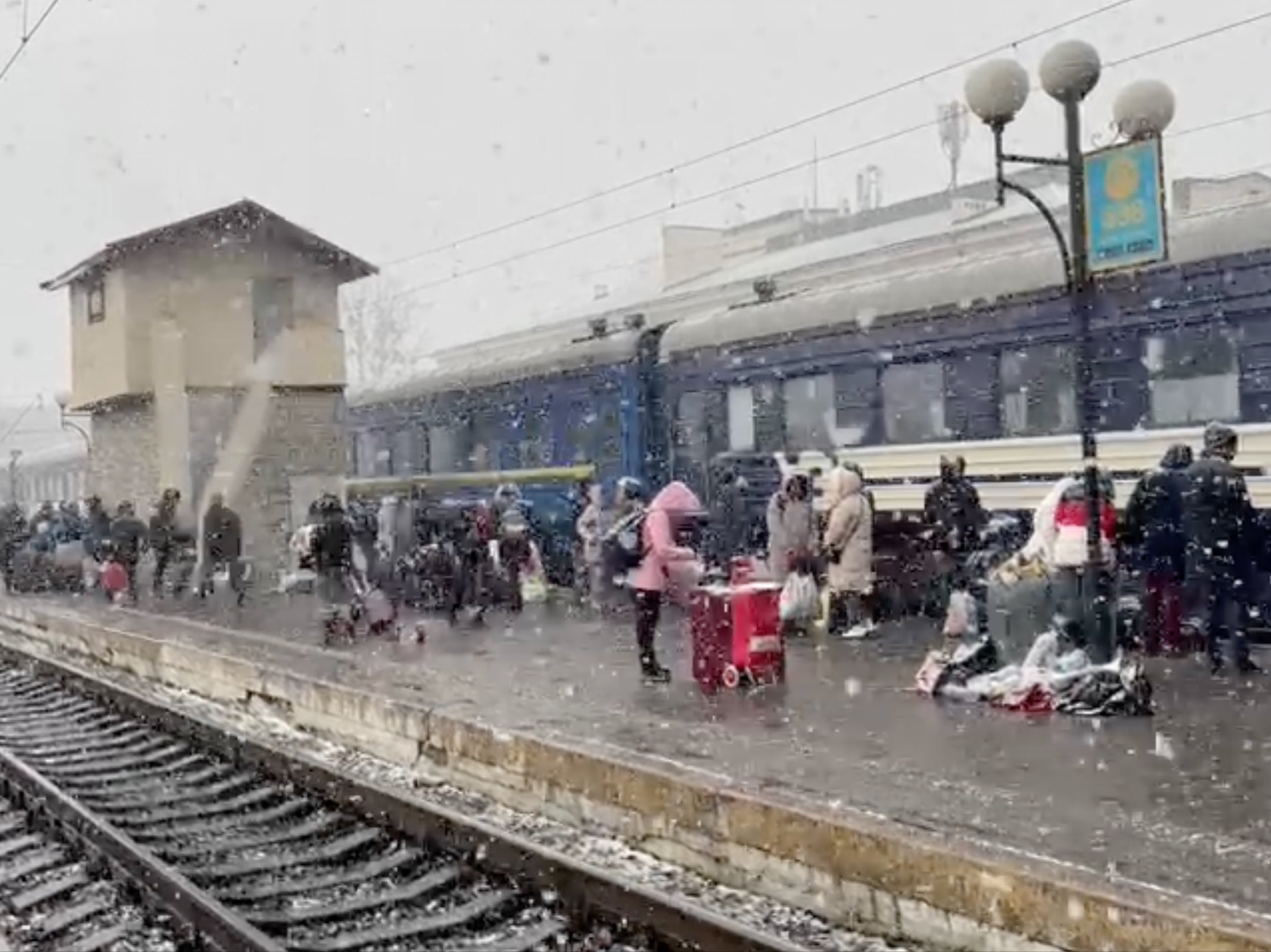 Snow falls as families wait together at a train station with their belongings