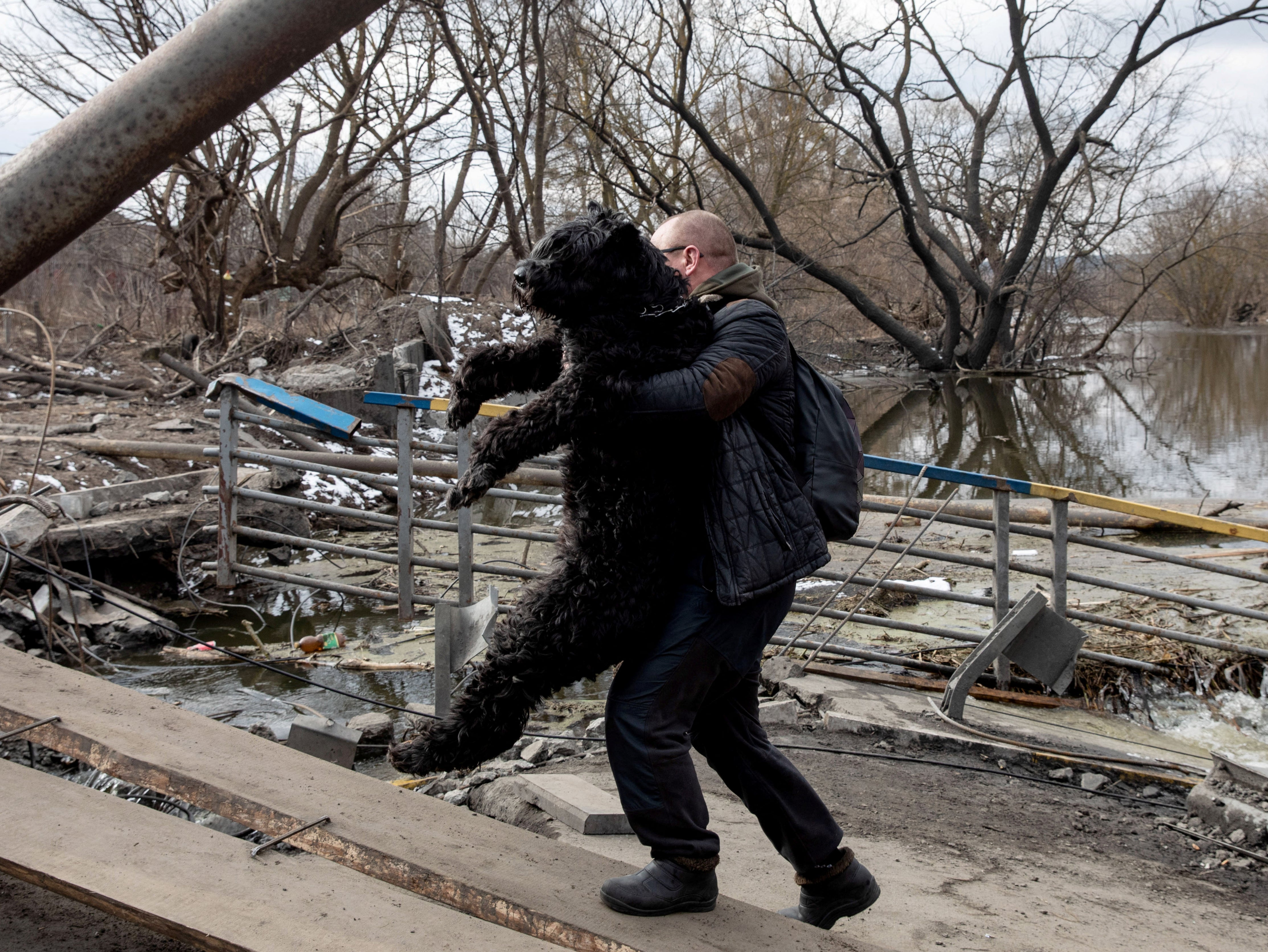 A man carries a dog as people flee near a destroyed bridge to cross the Irpin River