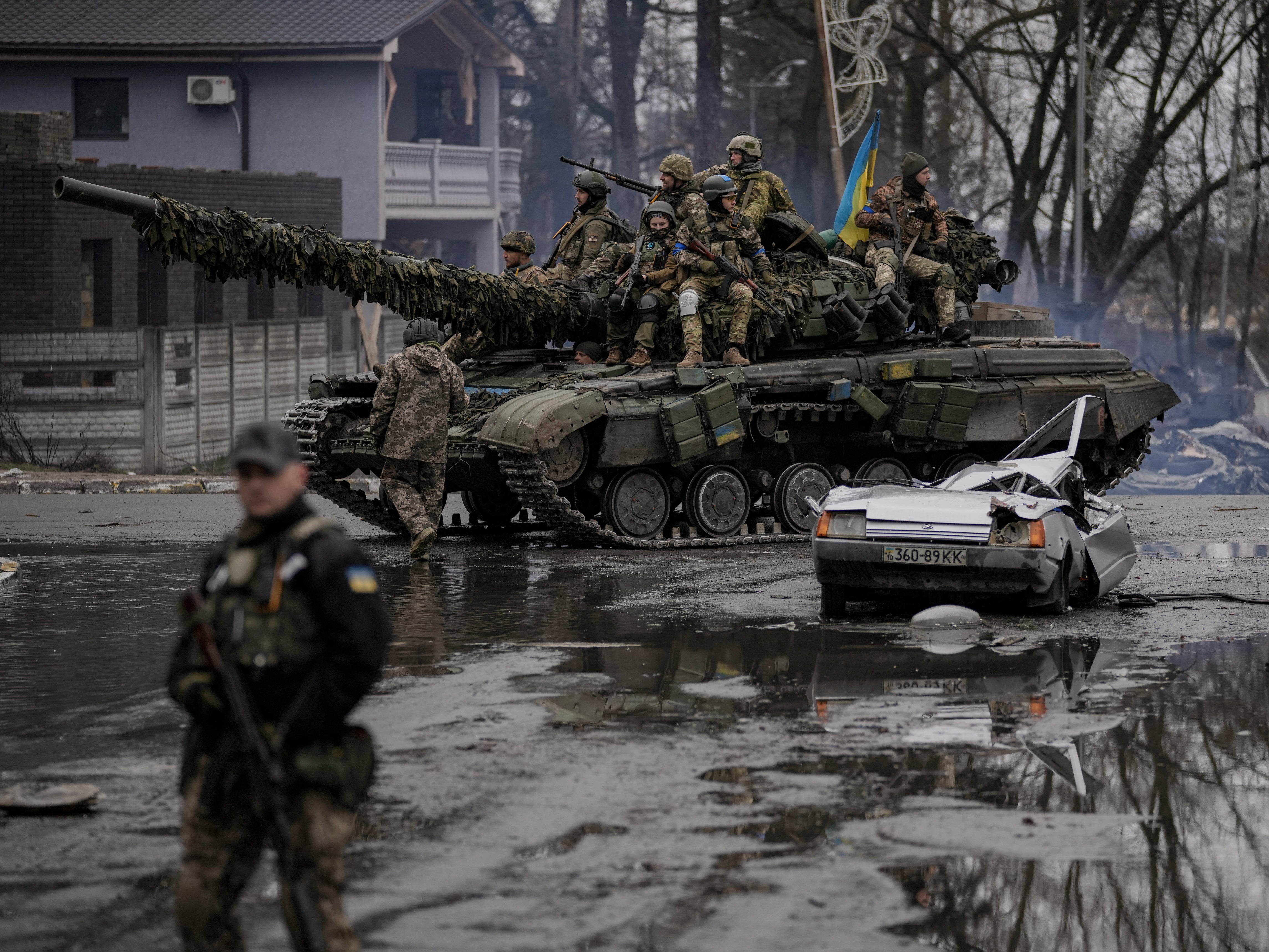 A man stands next to a civilian vehicle that was destroyed during fighting as Ukrainian servicemen ride on a tank vehicle outside Kyiv