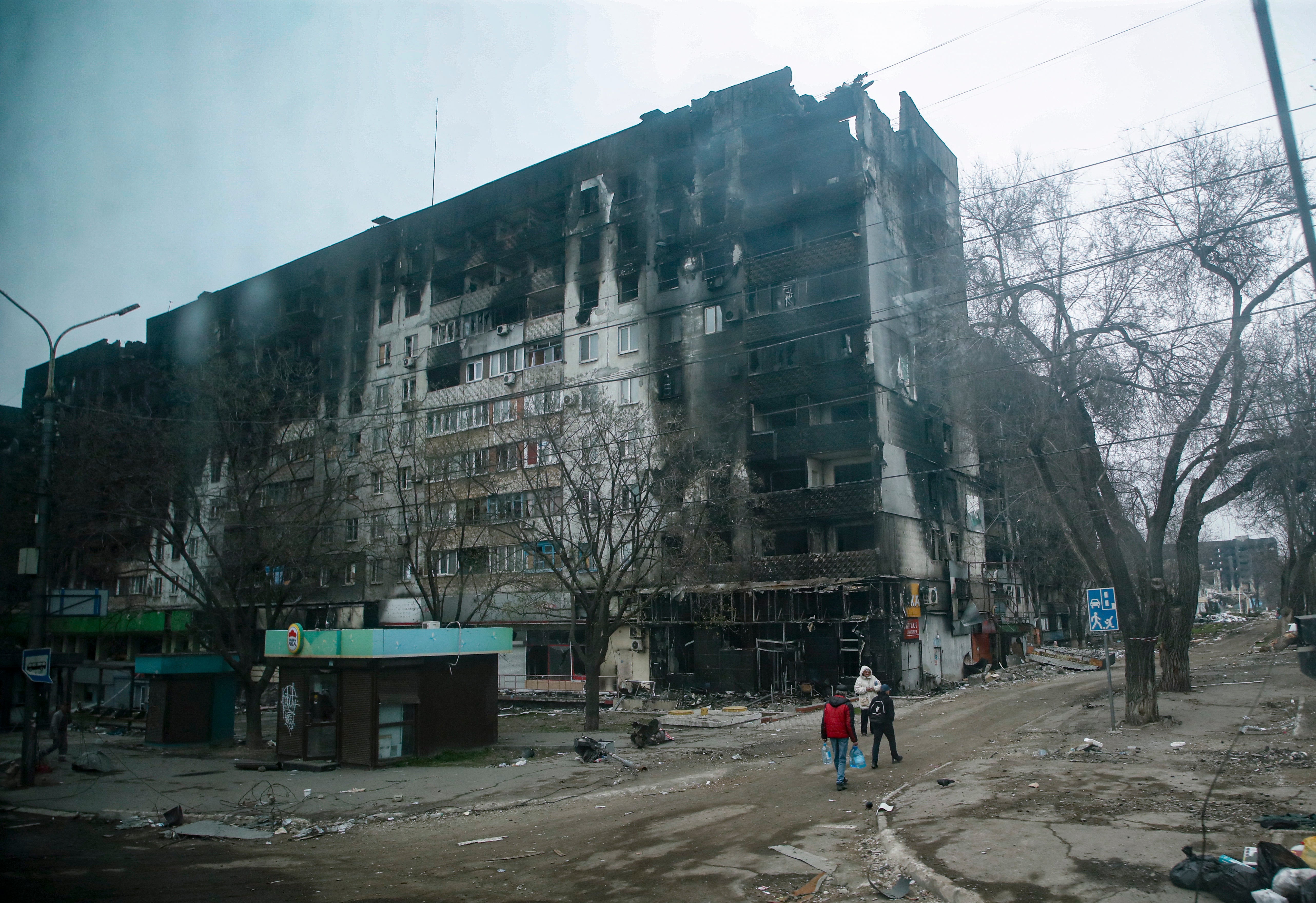 A picture taken during a visit to Mariupol organised by the Russian military shows local people carry water near a devastated and burned apartment building in Mariupol