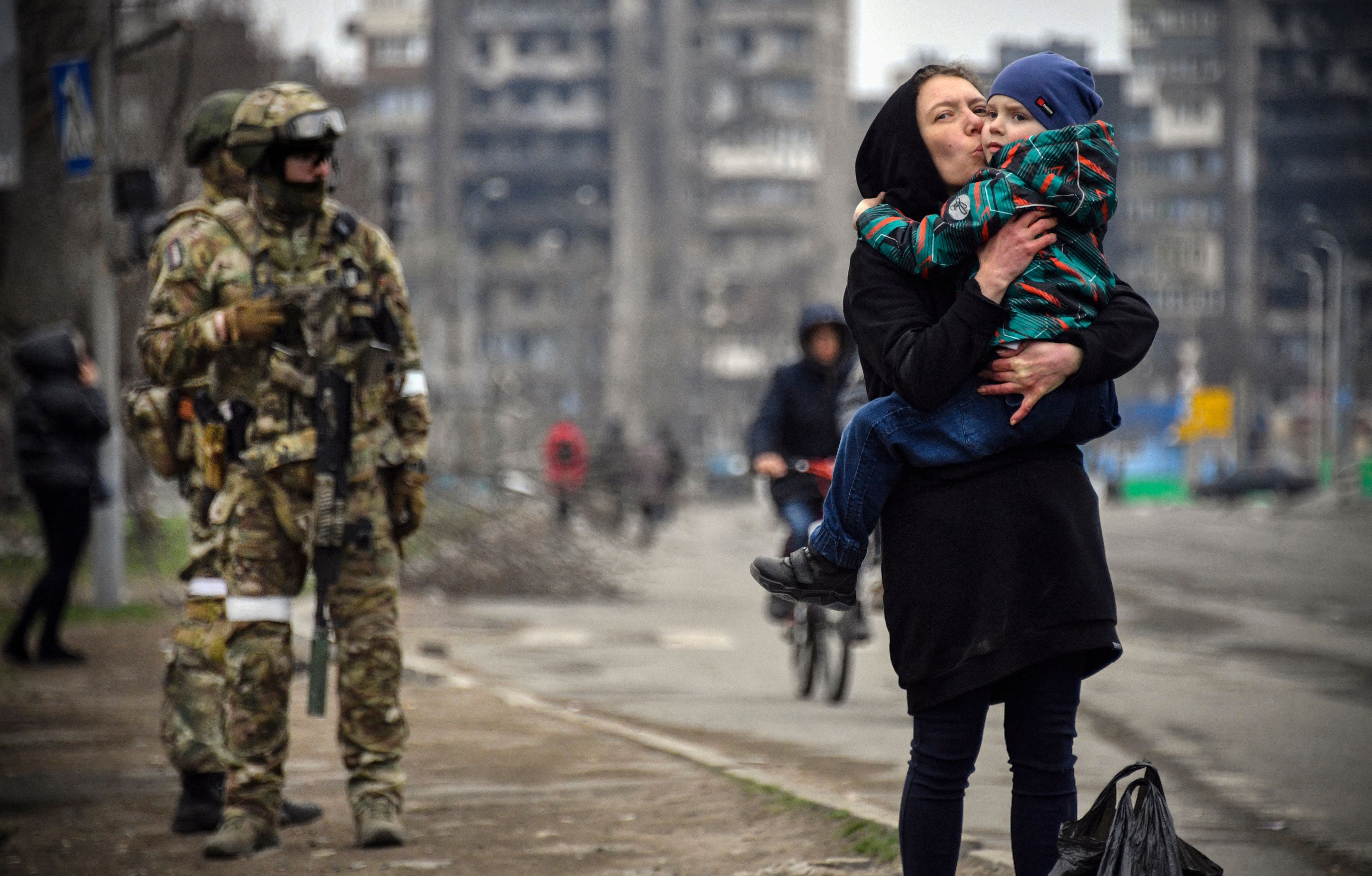 A civilian woman holds a child next to Russian soldiers in a Mariupol street