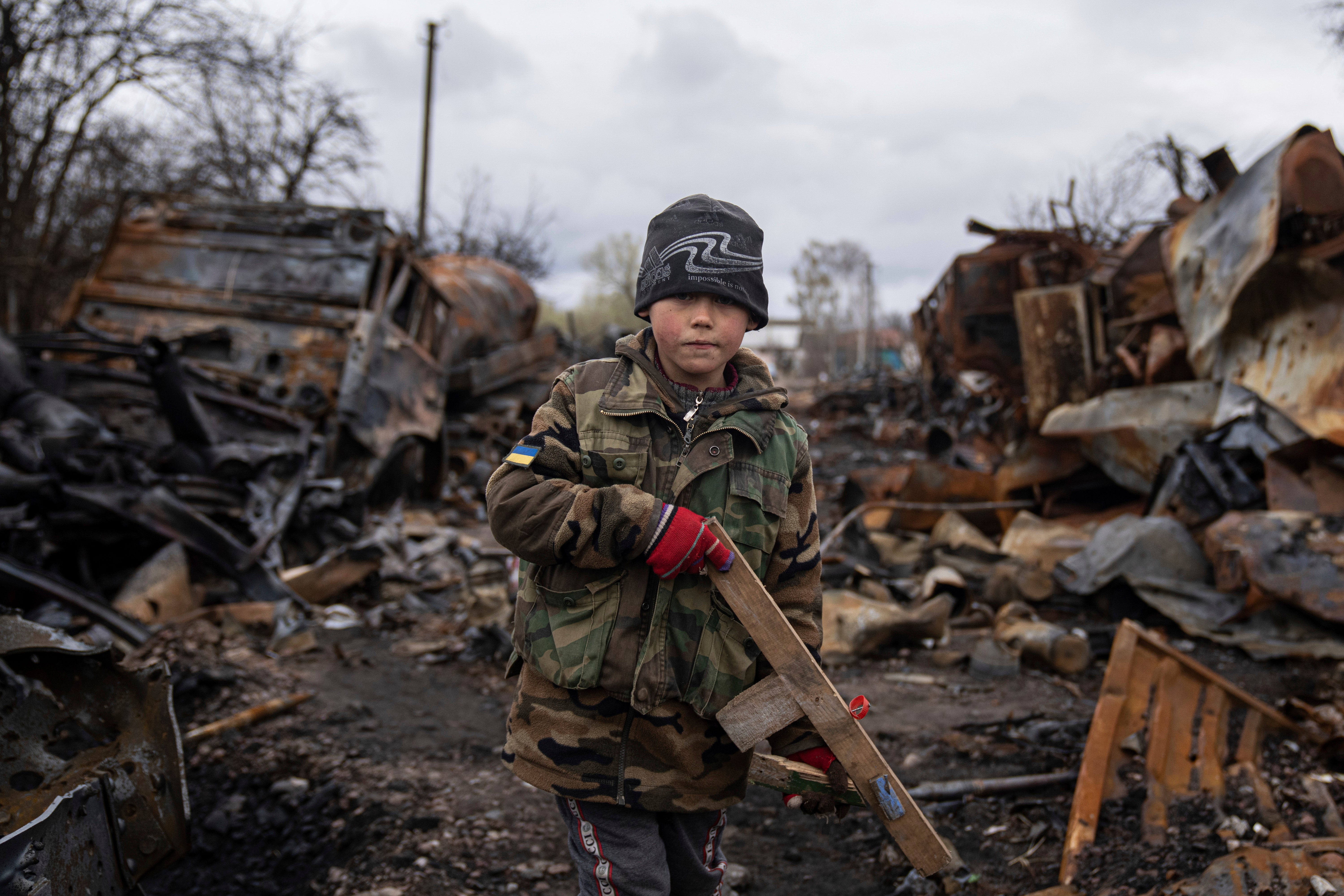 A boy holds a wooden toy rifle next to destroyed Russian military vehicles near Chernihiv