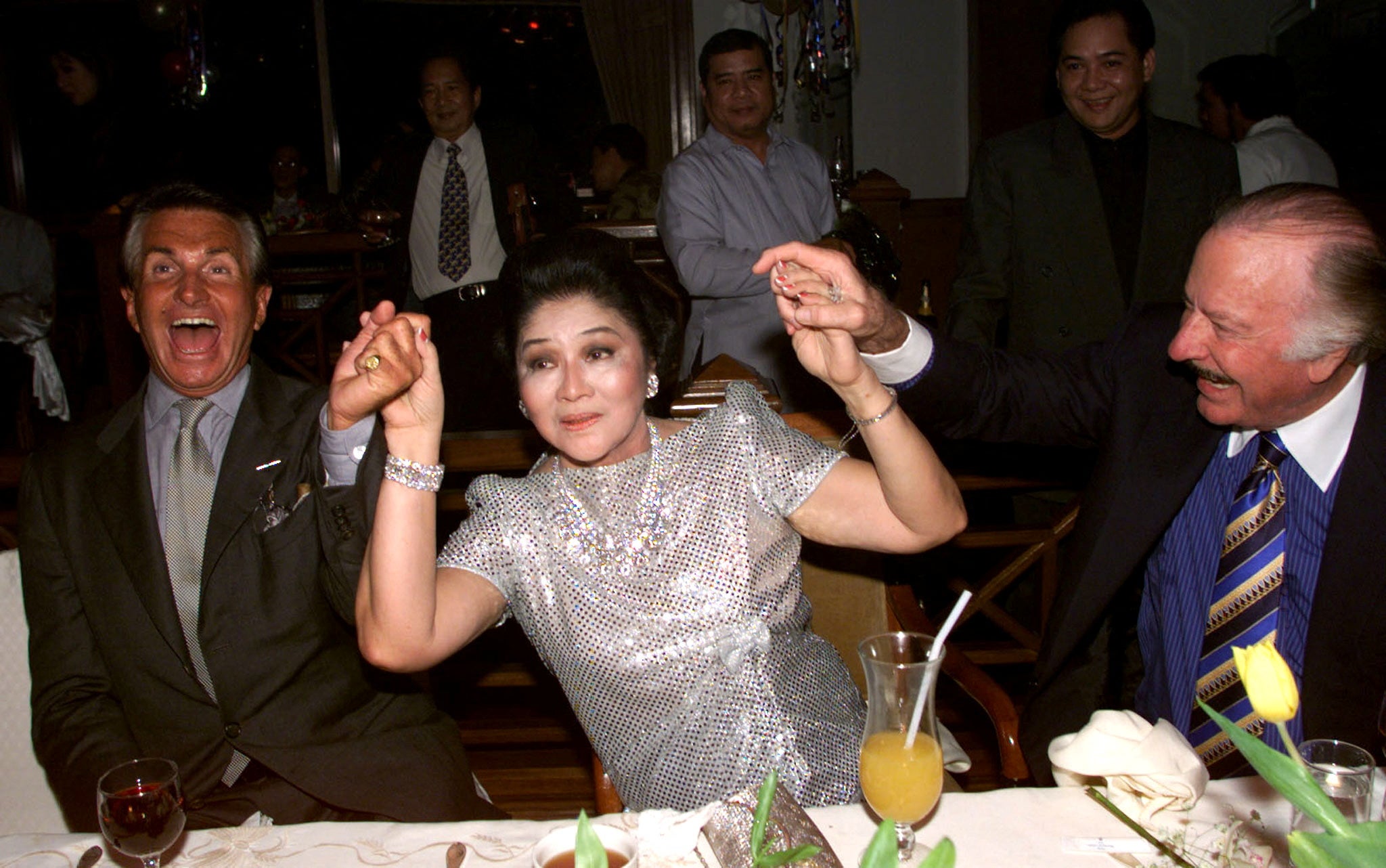 Hollywood actor George Hamilton and lawyer James Linn hold hands with Imelda Marcos as she celebrates her 70th birthday party