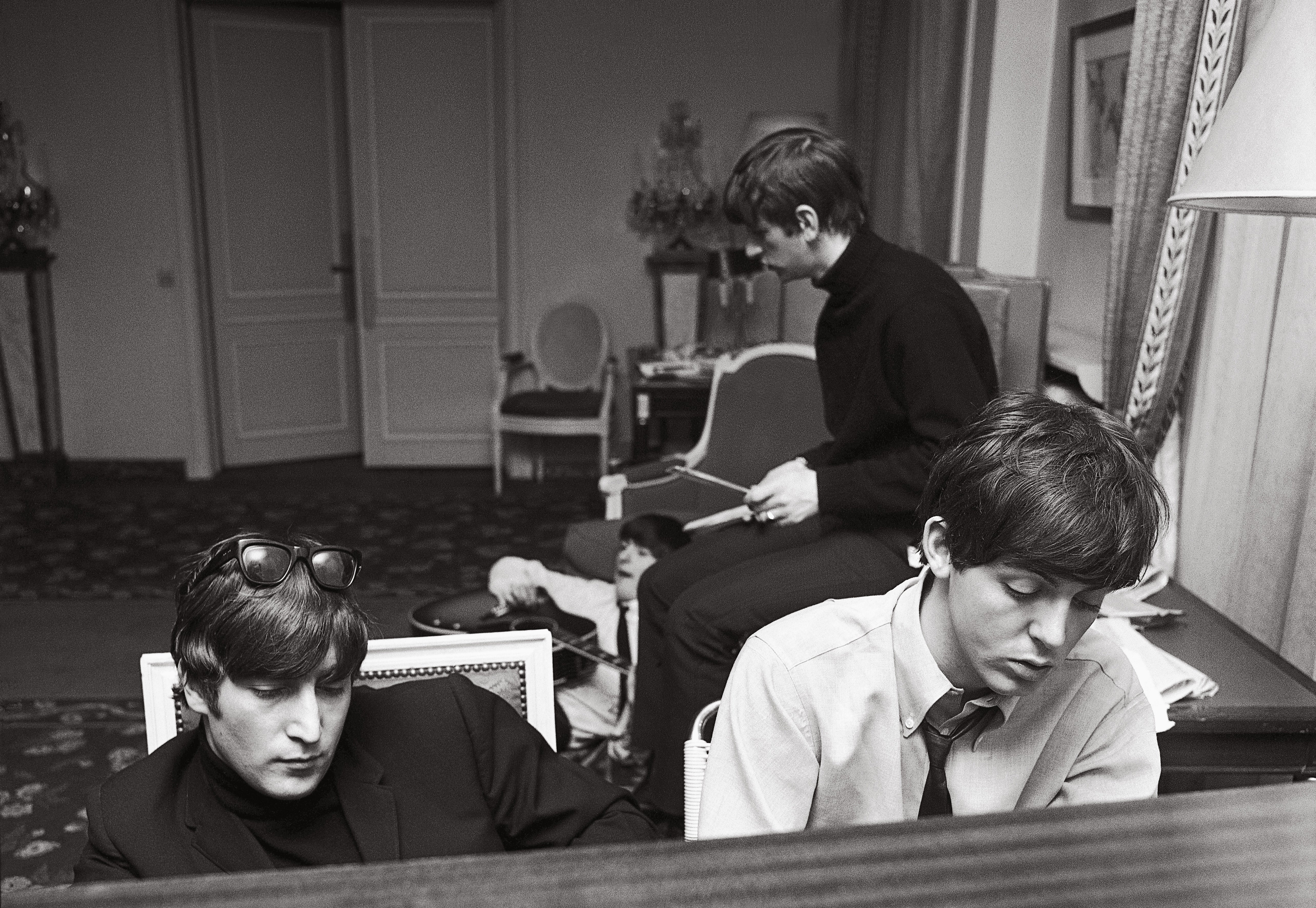 John and Paul deep in thought as they compose at the piano in their suite at the George V Hotel with George and Ringo in the background, January 1964