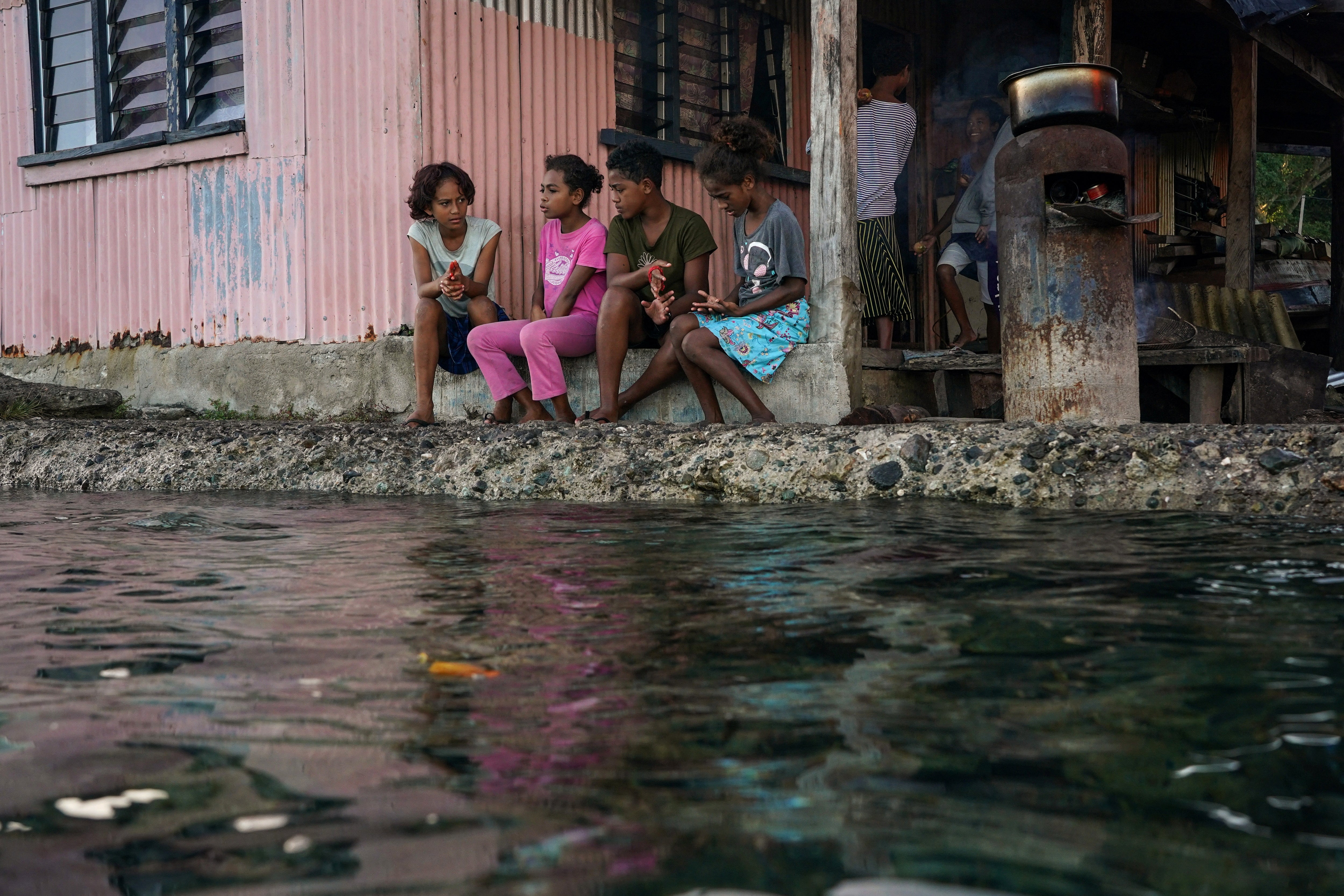 Village children pass the time in front of a home next to a flooding seawall at high tide in Serua village