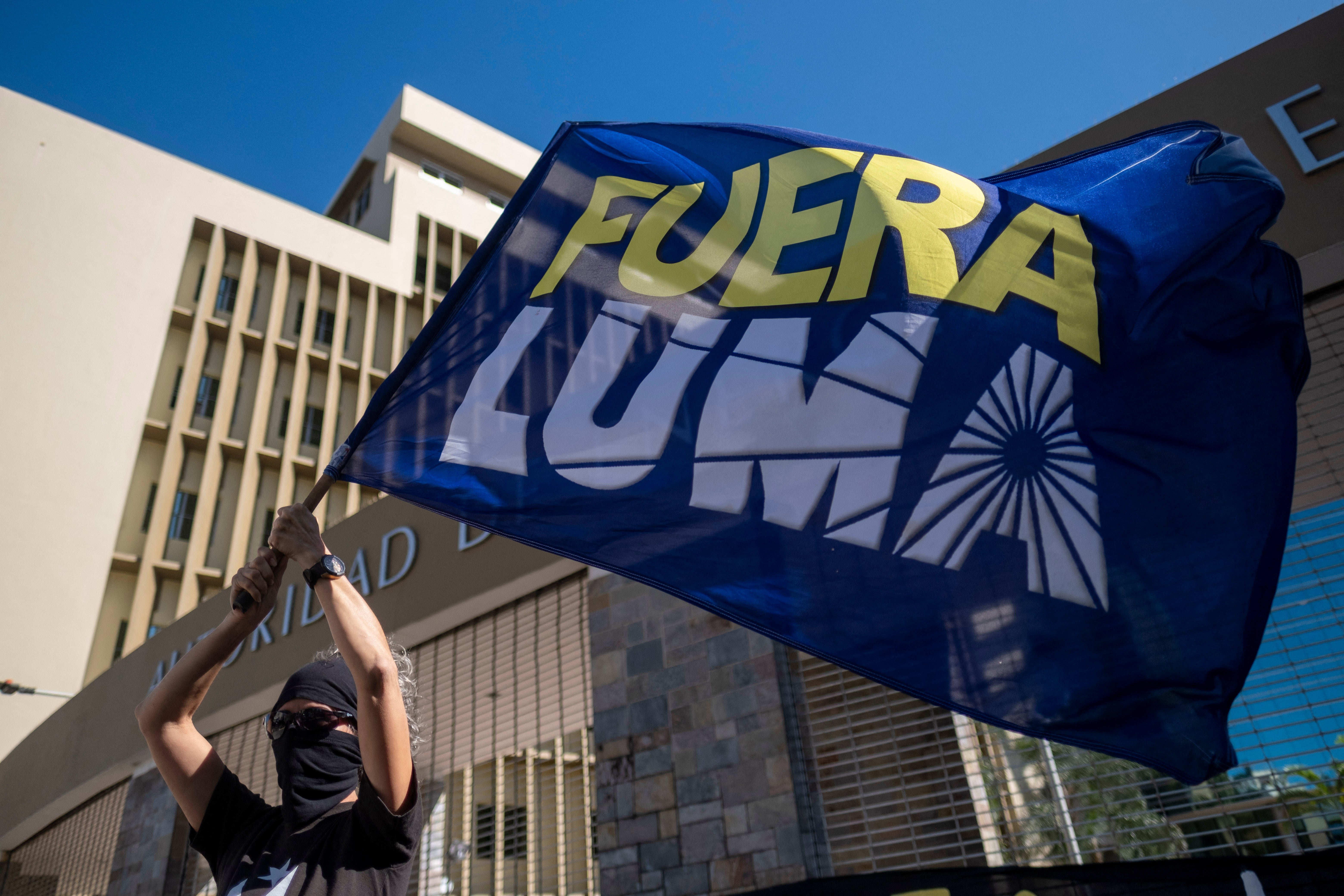 A woman waves a flag that reads in Spanish ‘LUMA Out’ during a protest in front of the headquarters of LUMA Energy