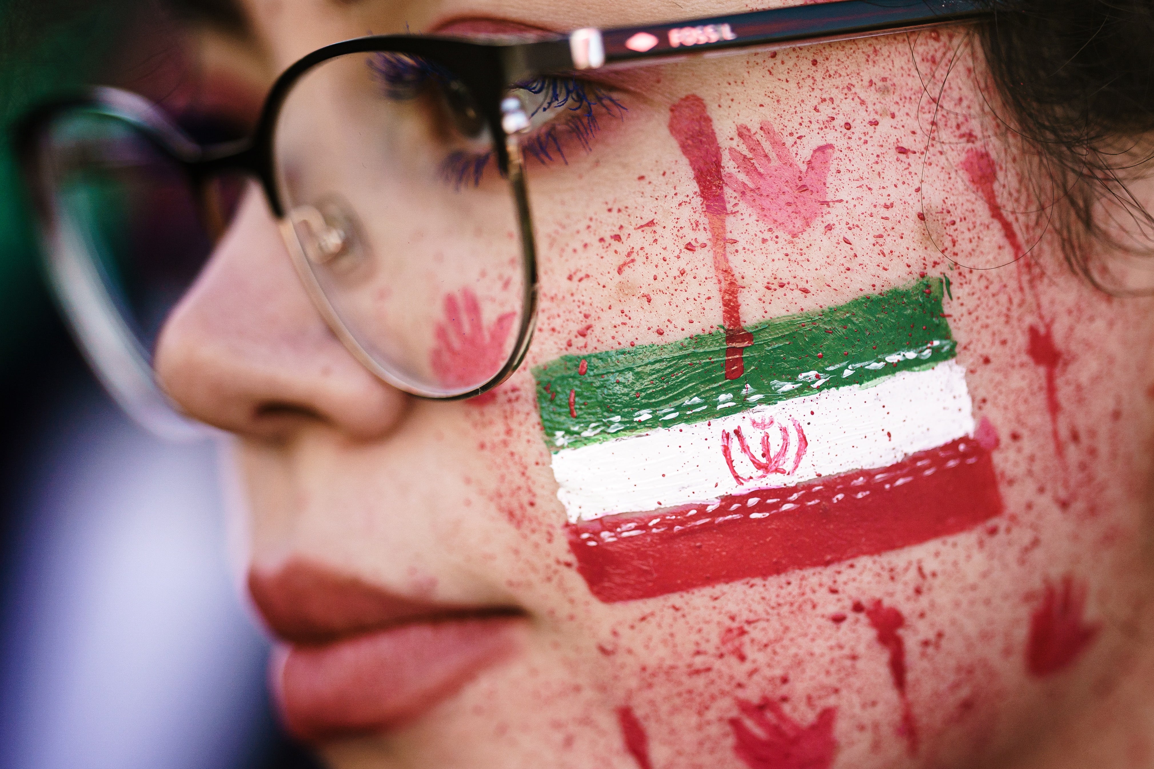 A protester shows red paint in her face resembling blood splatters and bloody handprints next to the national Iranian emblem