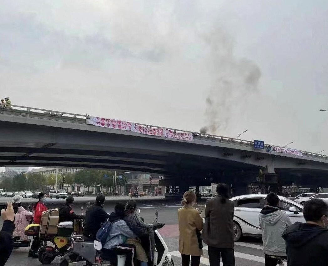 People watch while smoke rises as a banner with a protest message hangs off Sitong Bridge, Beijing