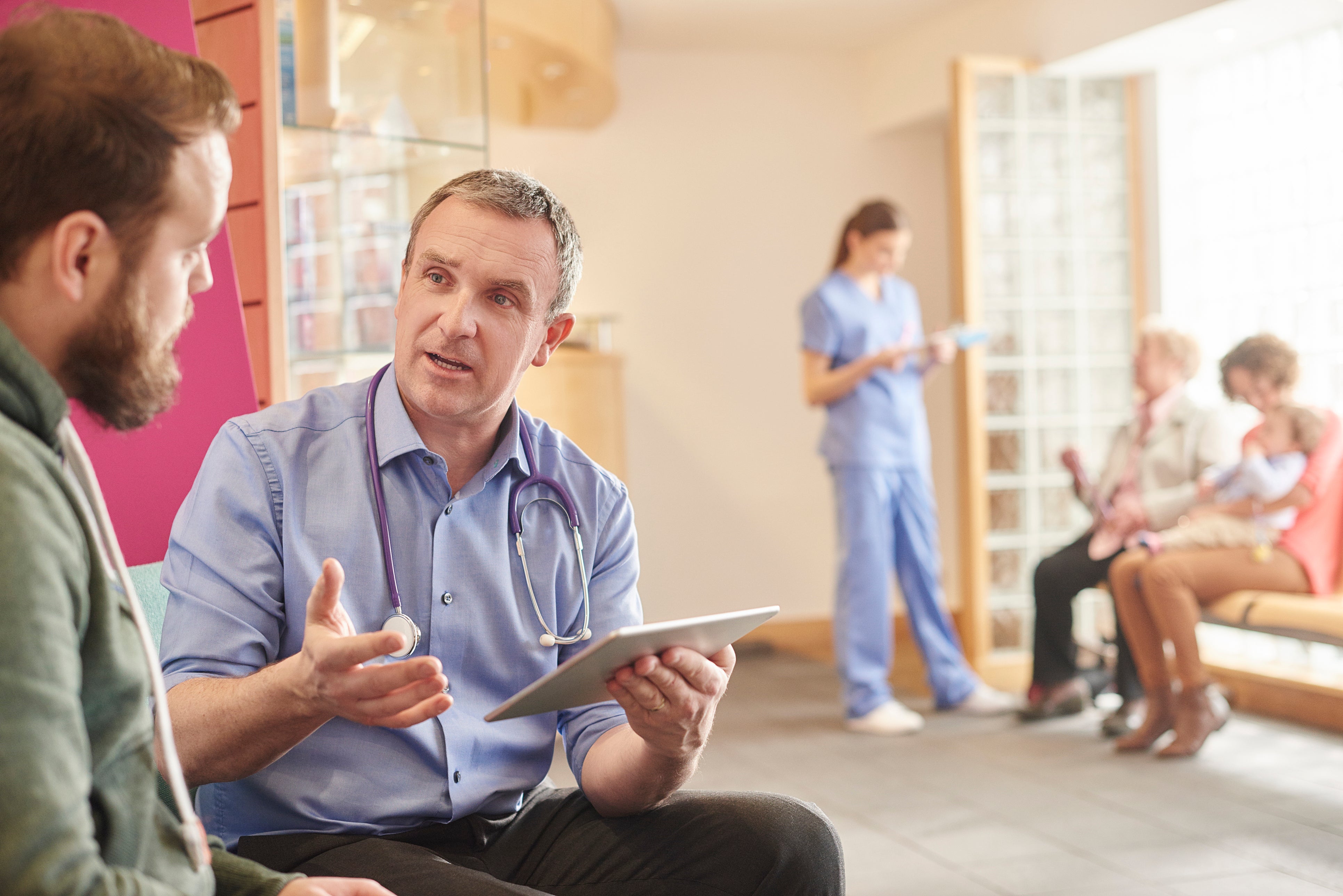 A mature male doctor sits next to a male patient in his waiting room and casually chats through a few things on his digital tablet . In the background a nurse chats to a mother and child and grandmother with her digital tablet .