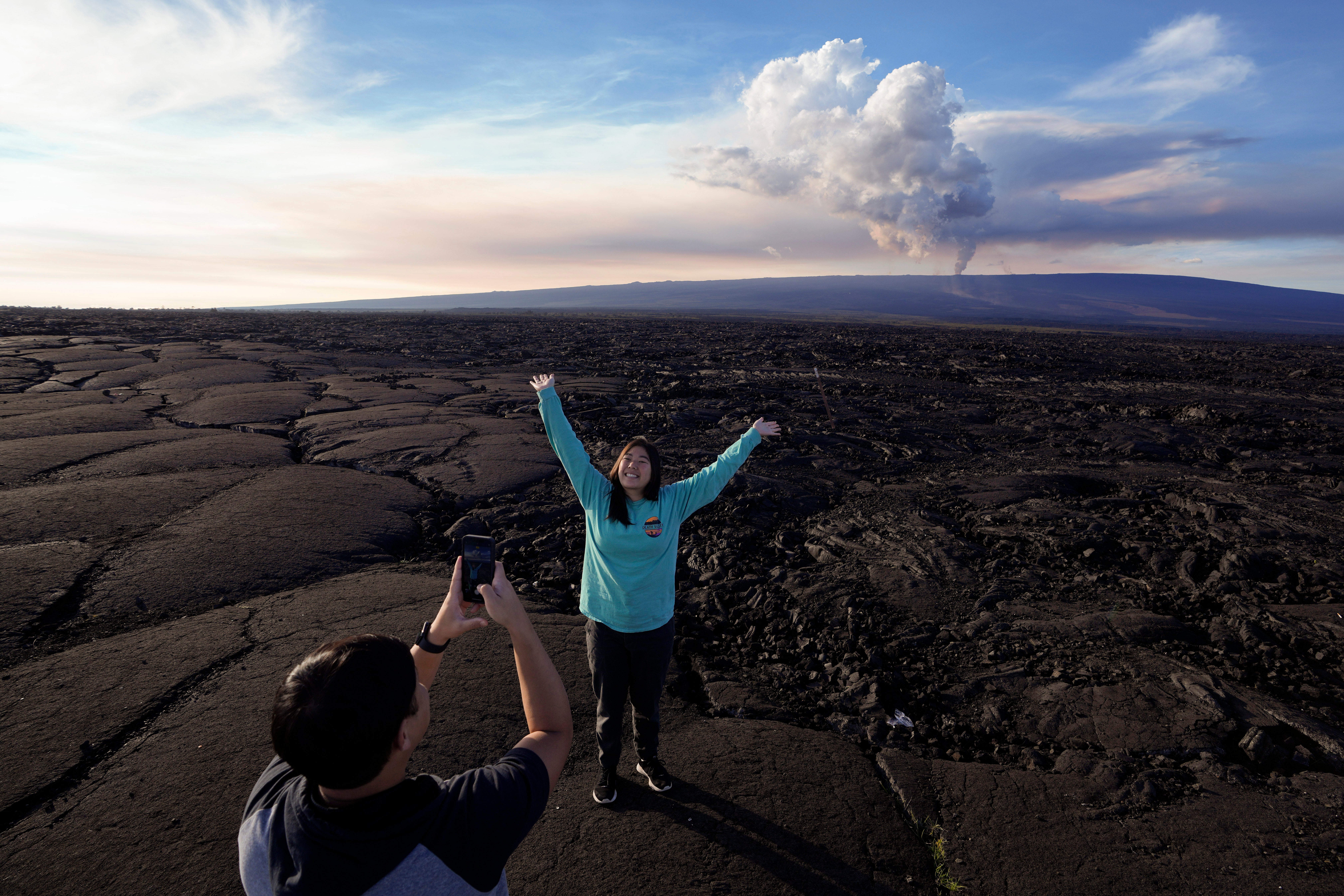 Kelly Ann Kobayashi raises her hands as she poses for a picture for Chad Saito, left, while standing on hardened lava rock from a previous eruption as the Mauna Loa volcano erupts, behind, Wednesday, Nov. 30, 2022, near Hilo, Hawaii