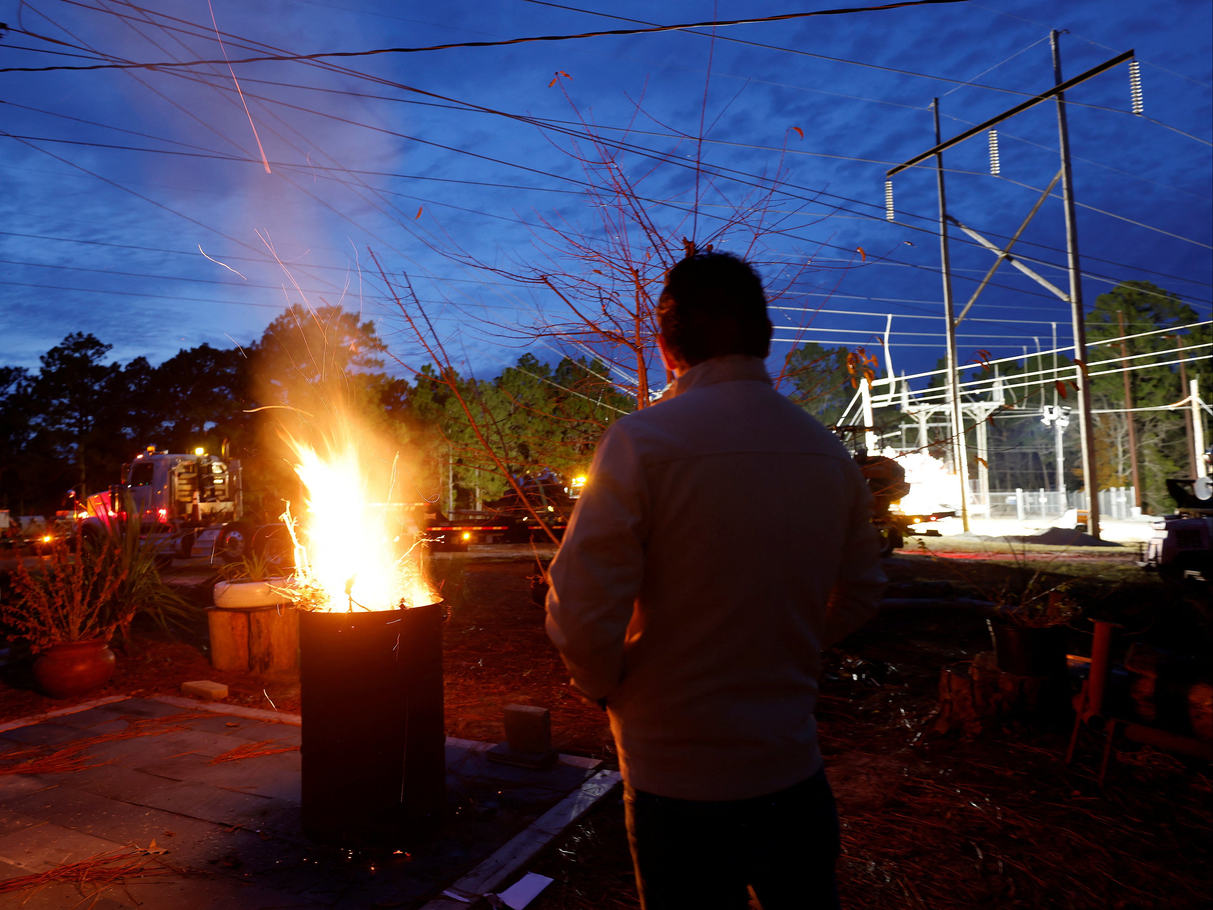 A man warms himself in front of a makeshift fire as he watches Duke Energy personnel work to restore power at a crippled electrical substation in Moore County, North Carolina