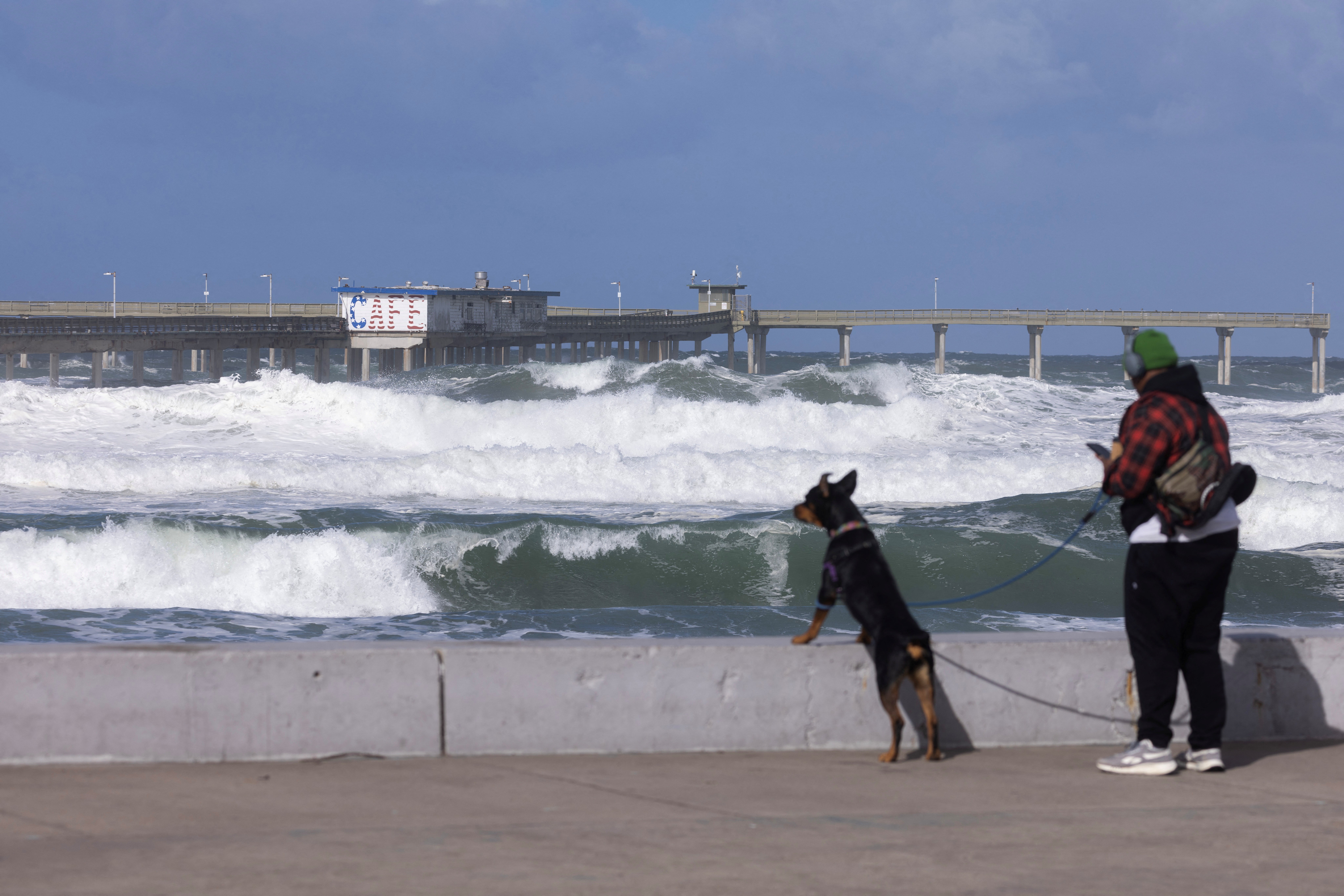 A person walking their dog stands next to the ocean as large wind-driven waves hit the pier in the Ocean Beach neighborhood of San Diego, California on February 22 (REUTERS/Mike Blake)