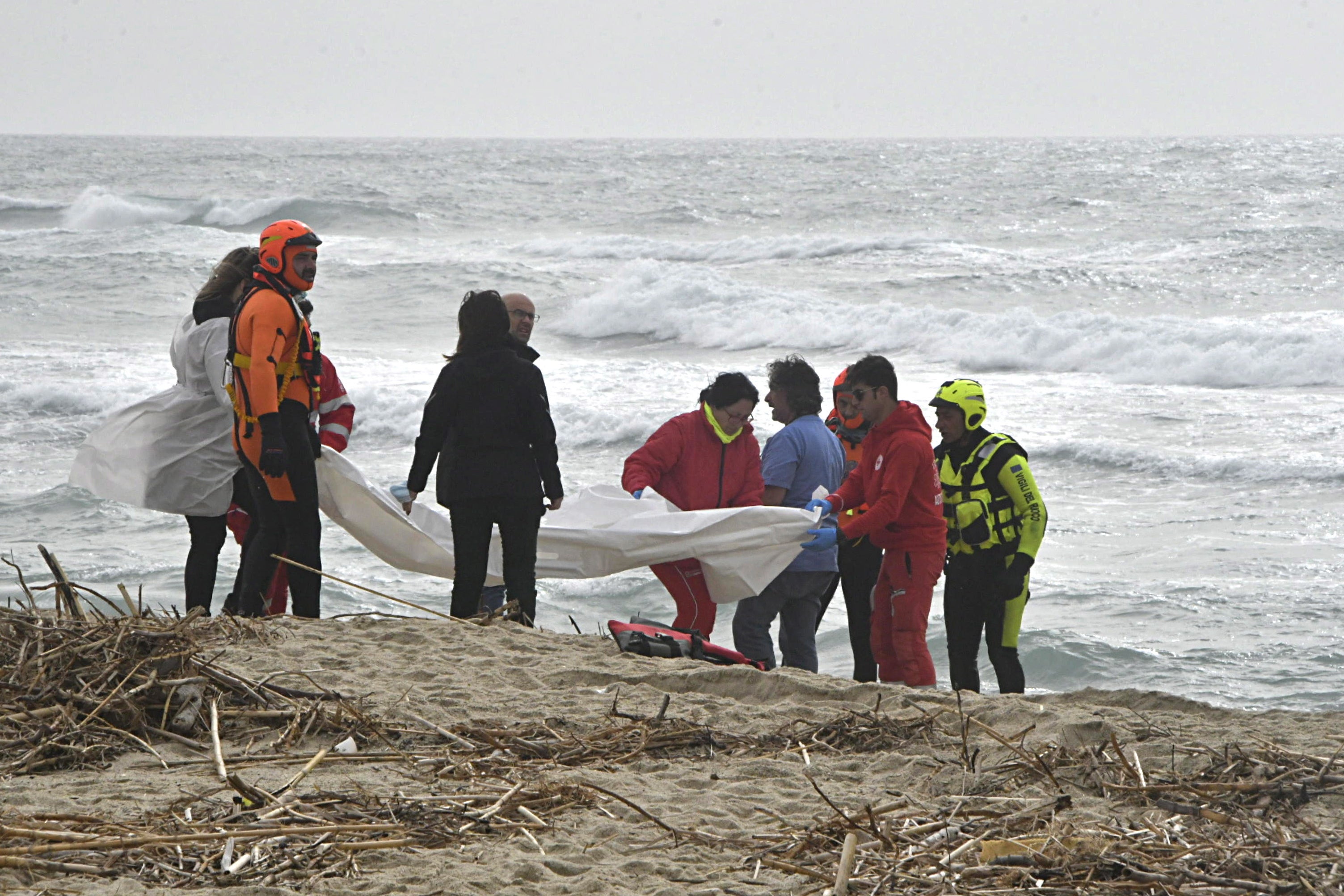 Italian firefighters and Red Cross personnel gather on the beach, surrounded by debris