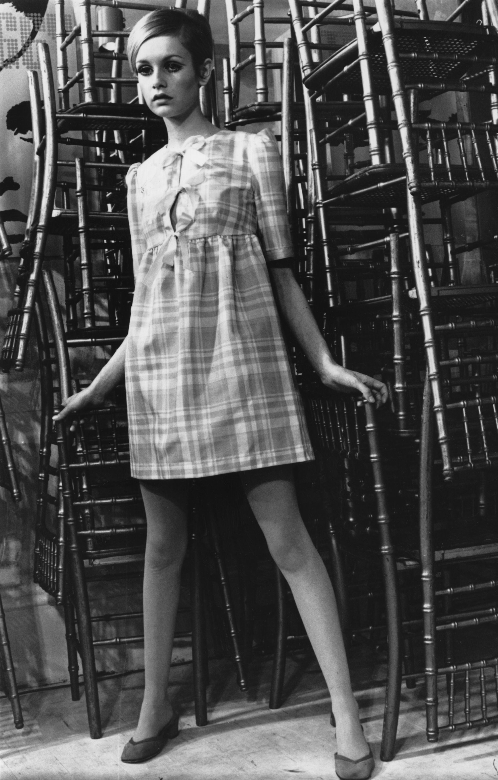 Twiggy, originally Lesley Hornby, poses in font of a stack of chairs.