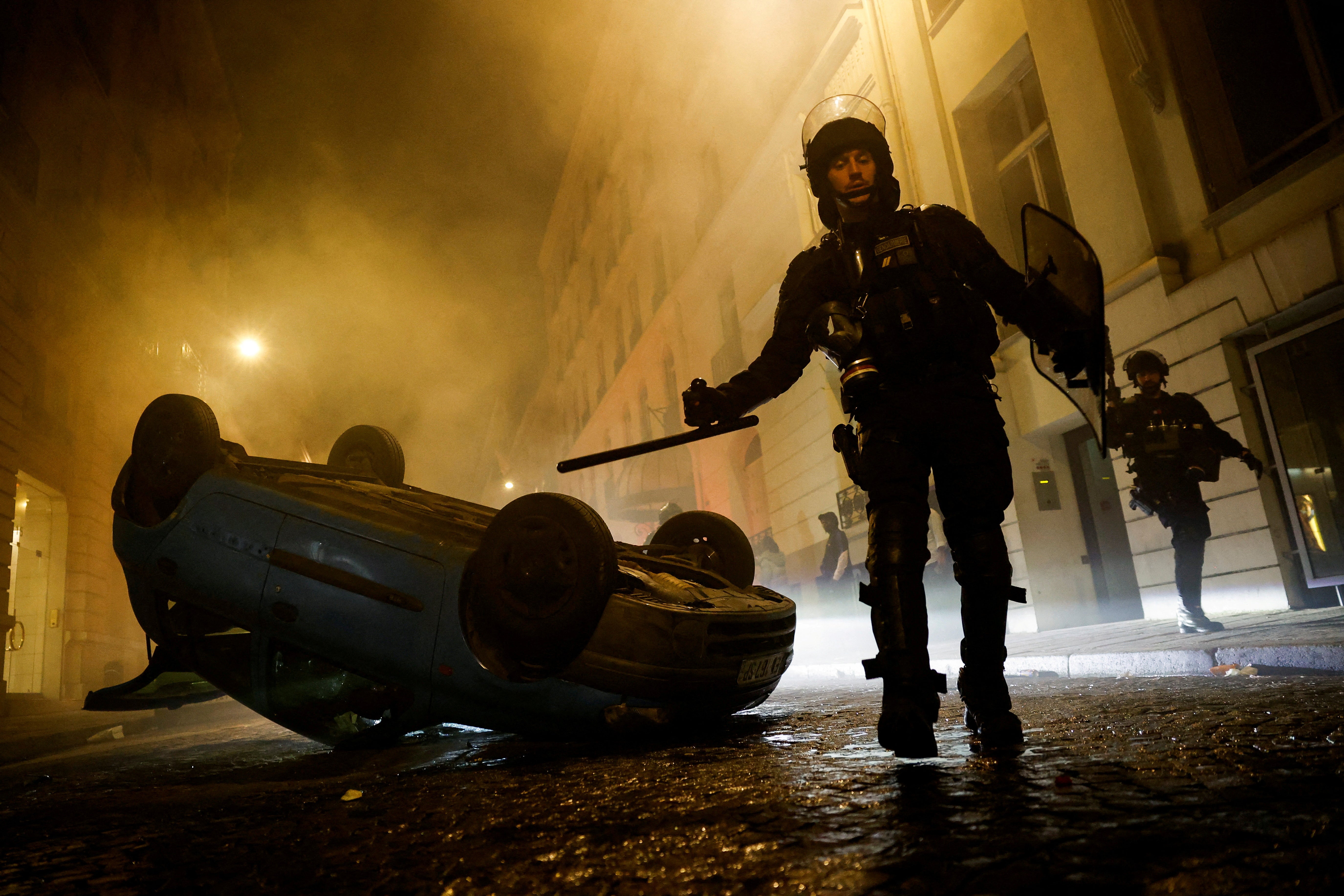 French riot police officers walk next to a vehicle upside down