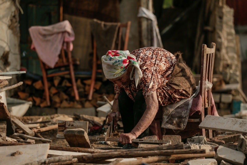 Nina cuts wood while sitting on a chair amid the remains of her house