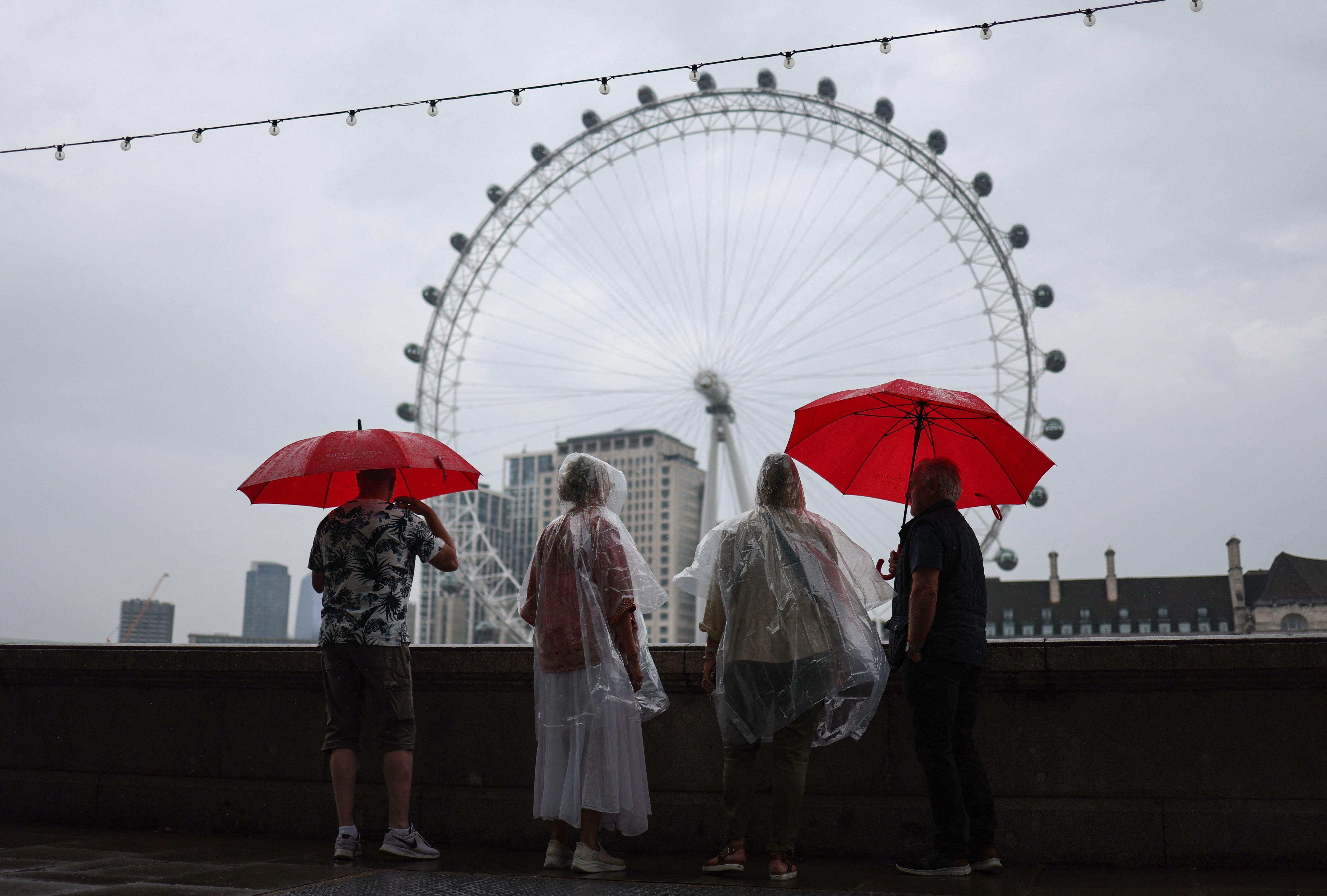 Pedestrians shelter from the rain beneath umbrellas while looking at the London landmark, the London Eye, from Embankment by the River Thames, in central London