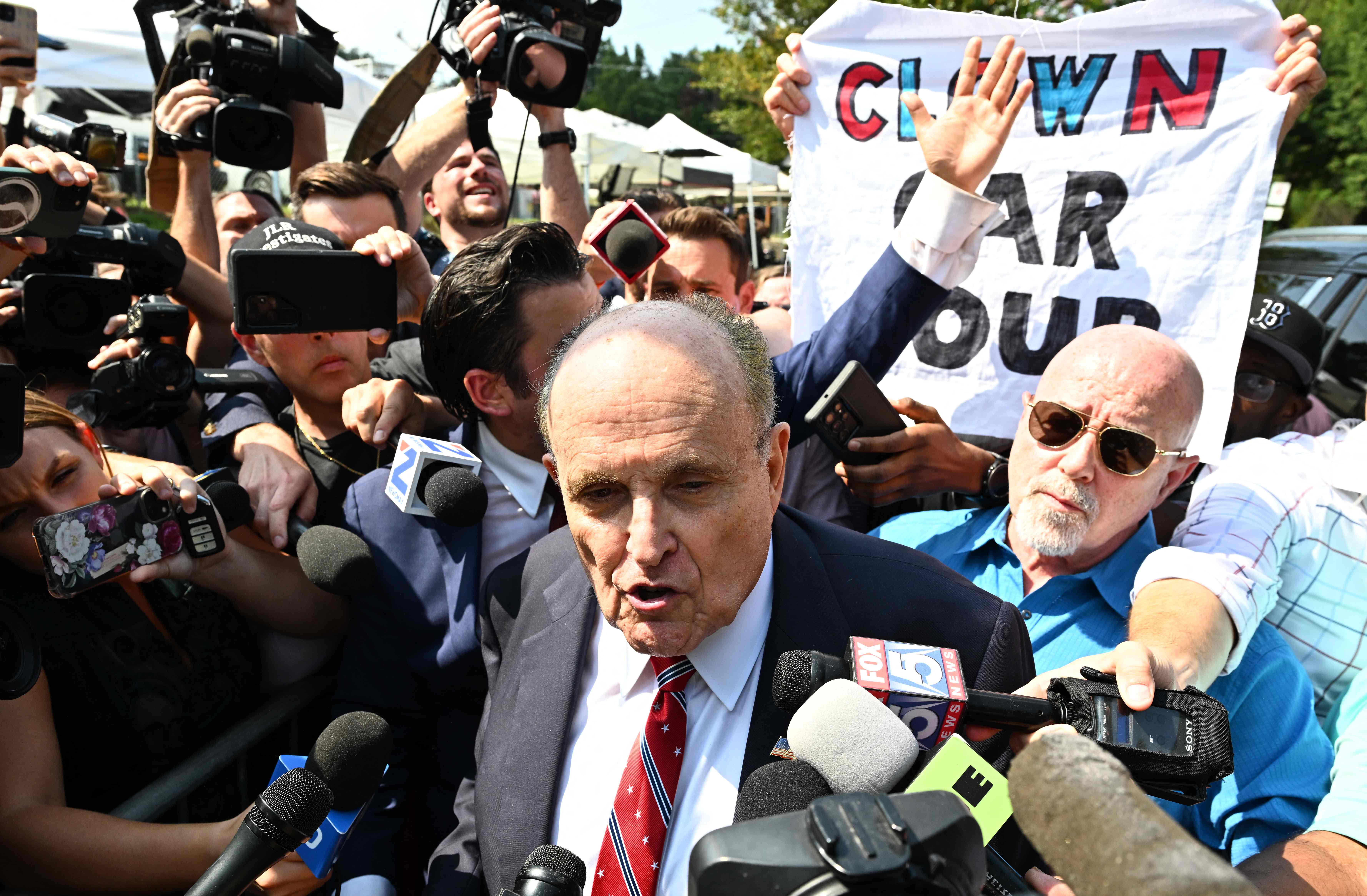 A person holds a sign reading ‘Clown Car Coup’ as Rudy Giuliani speaks to the media after being booked at the Fulton County Jail in Atlanta, Georgia