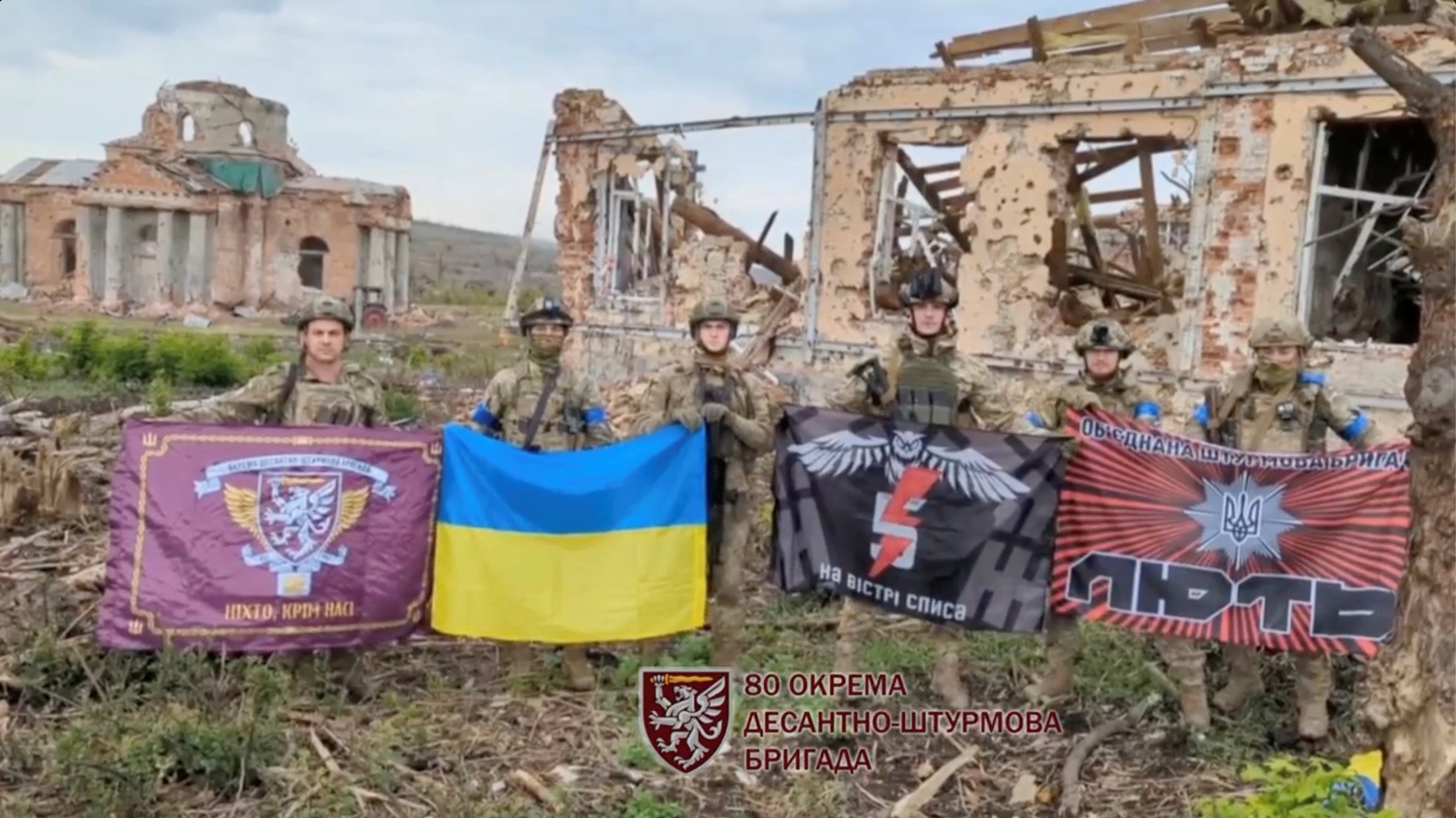 Soldiers hold flags as they speak in front of destroyed buildings in Klishchiivka