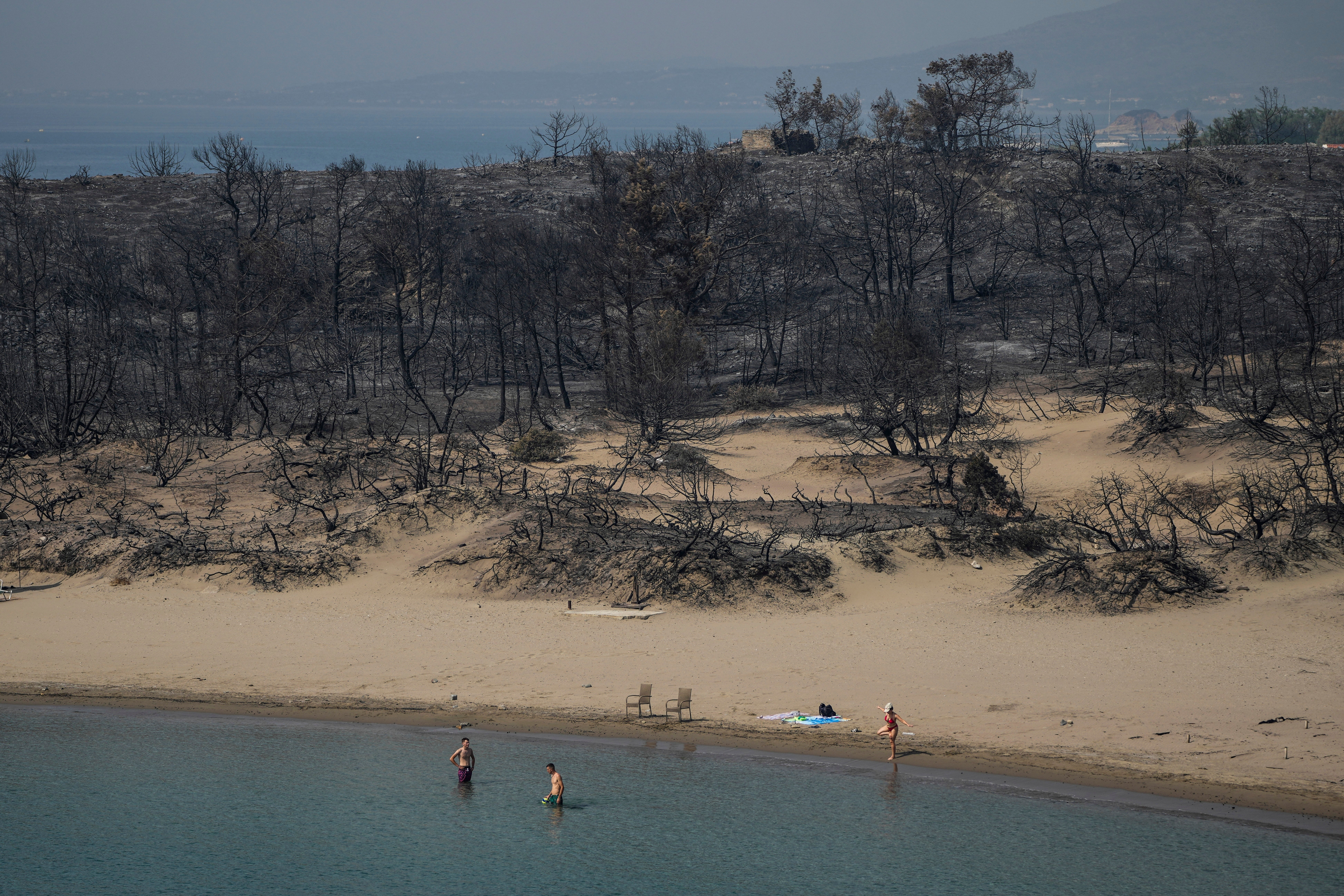 People gather on a beach in front of burnt forest near Gennadi village, on the Aegean Sea island of Rhodes, southeastern Greece, on Thursday, July 27, 2023