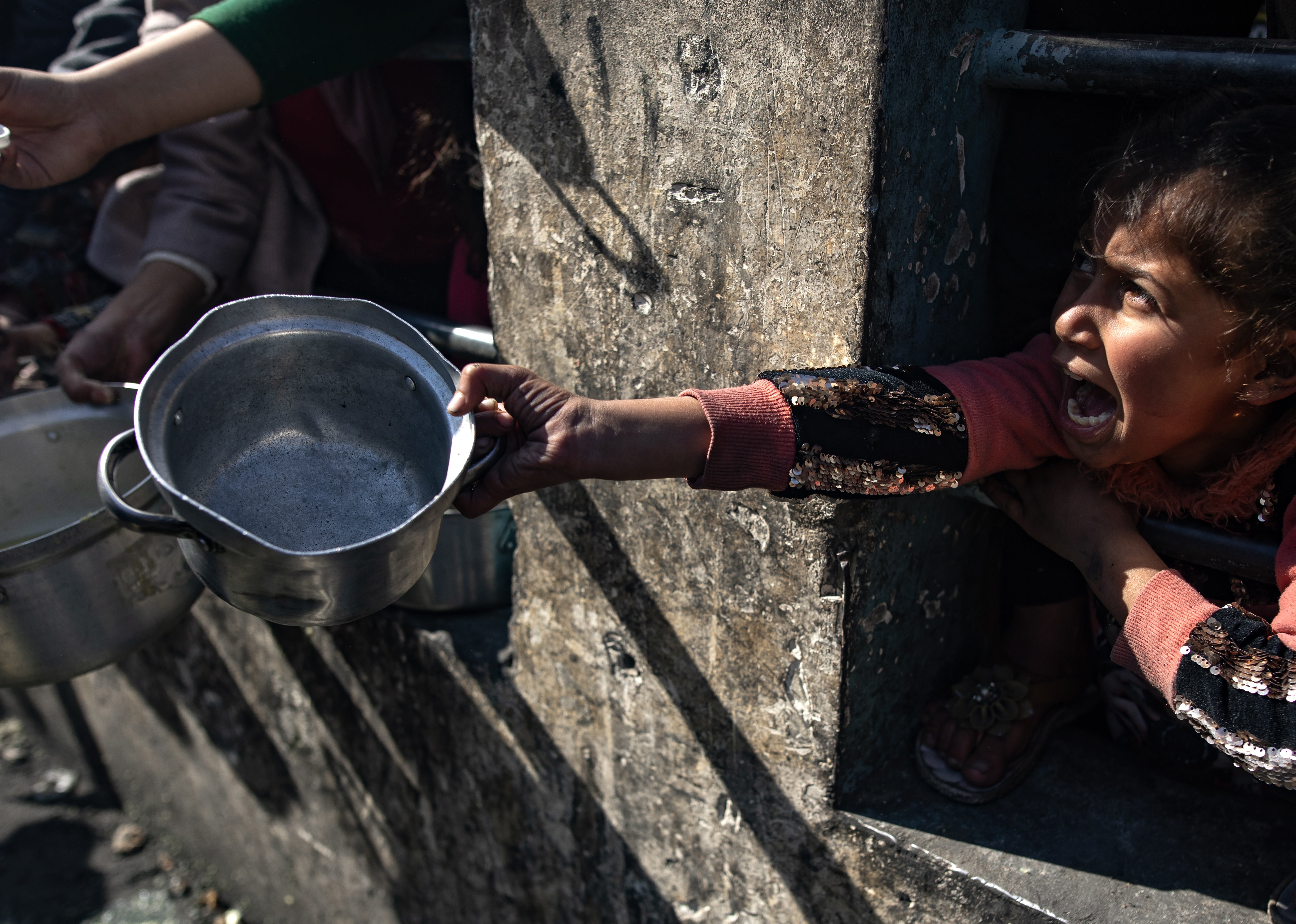 A displaced Palestinian child holds up an empty pot as she waits with others to receive food aid