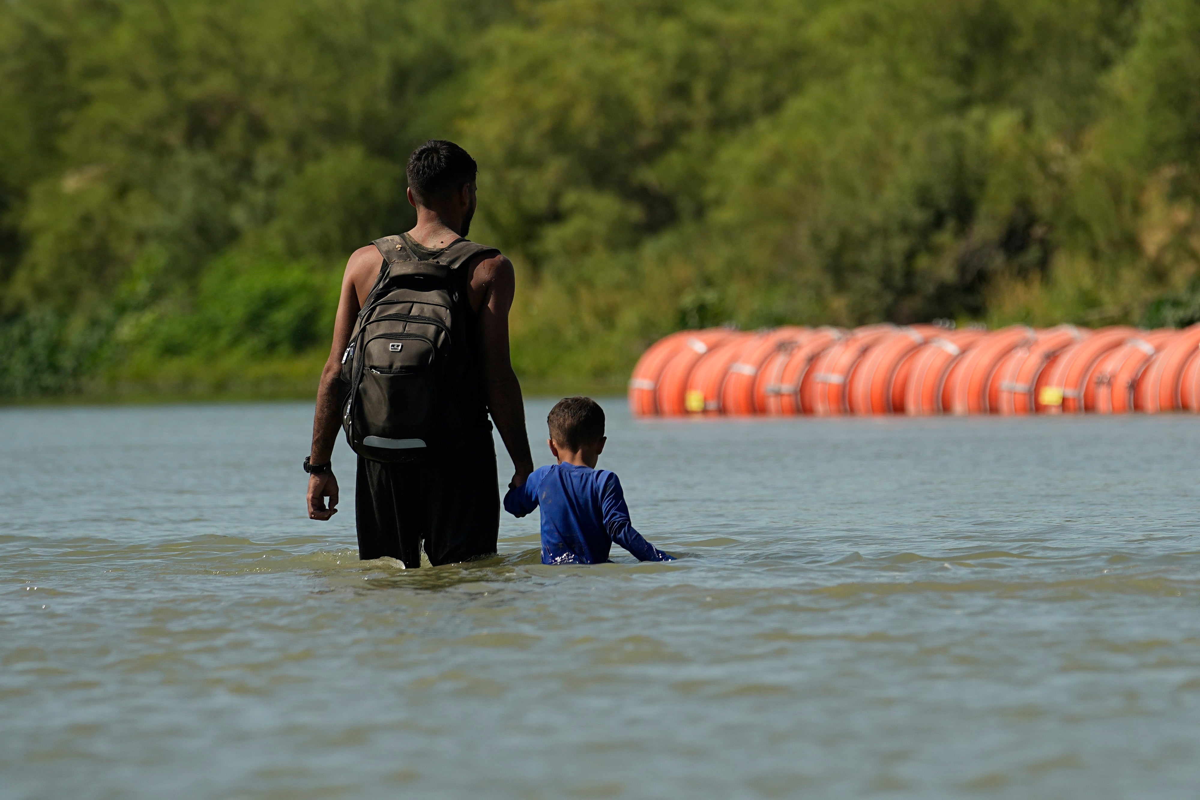 Migrants walk past large buoys being used as a floating border barrier on the Rio Grande, Aug. 1, 2023, in Eagle Pass, Texas. A US appeals court overturned a previous ruling requiring the barrier to be removed on July 31, 2024