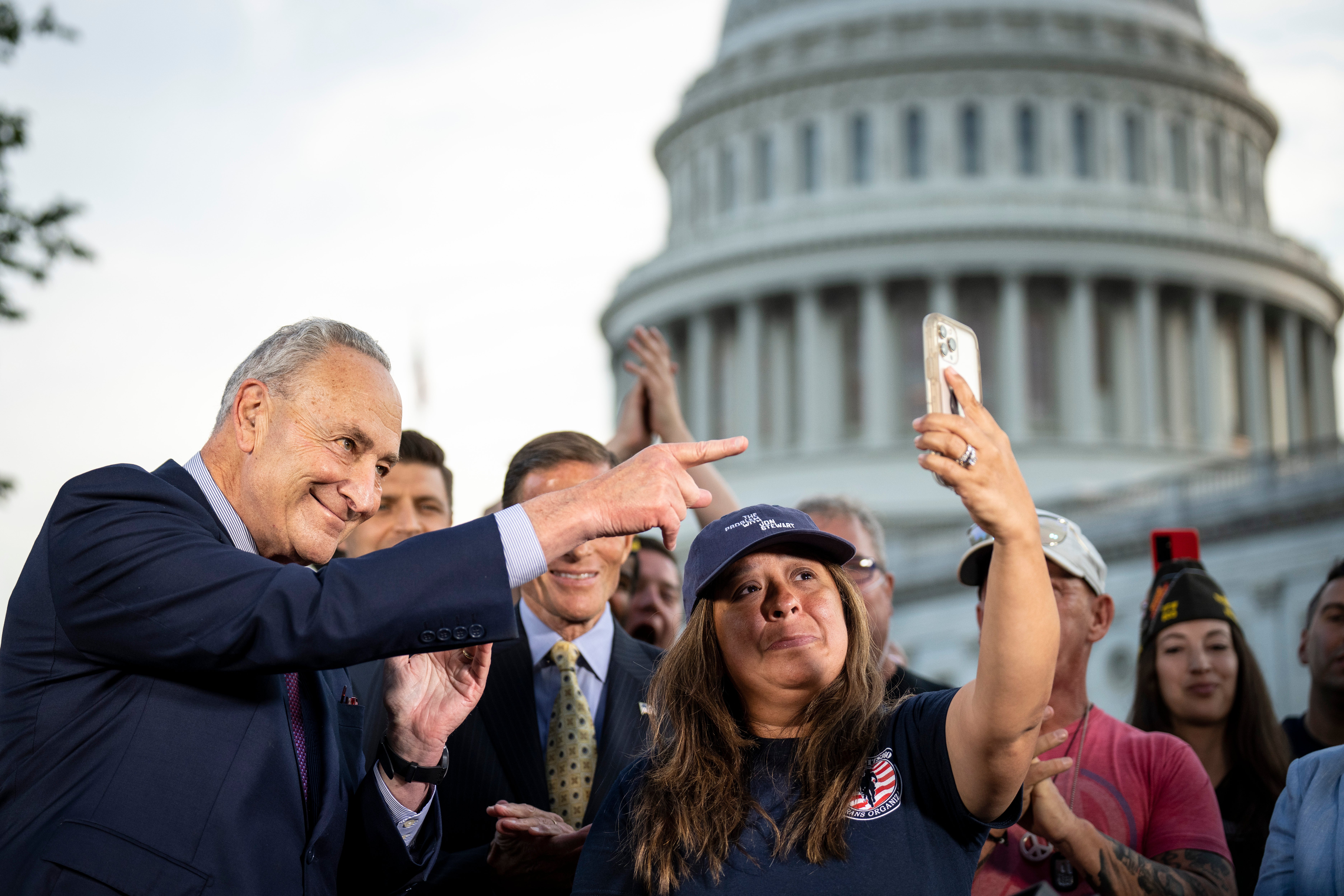 Senate Majority Leader Chuck Schumer looks on as Rosie Torres holds up her husband Le Roy on her phone in August 2022 as they celebrate the bill’s passage