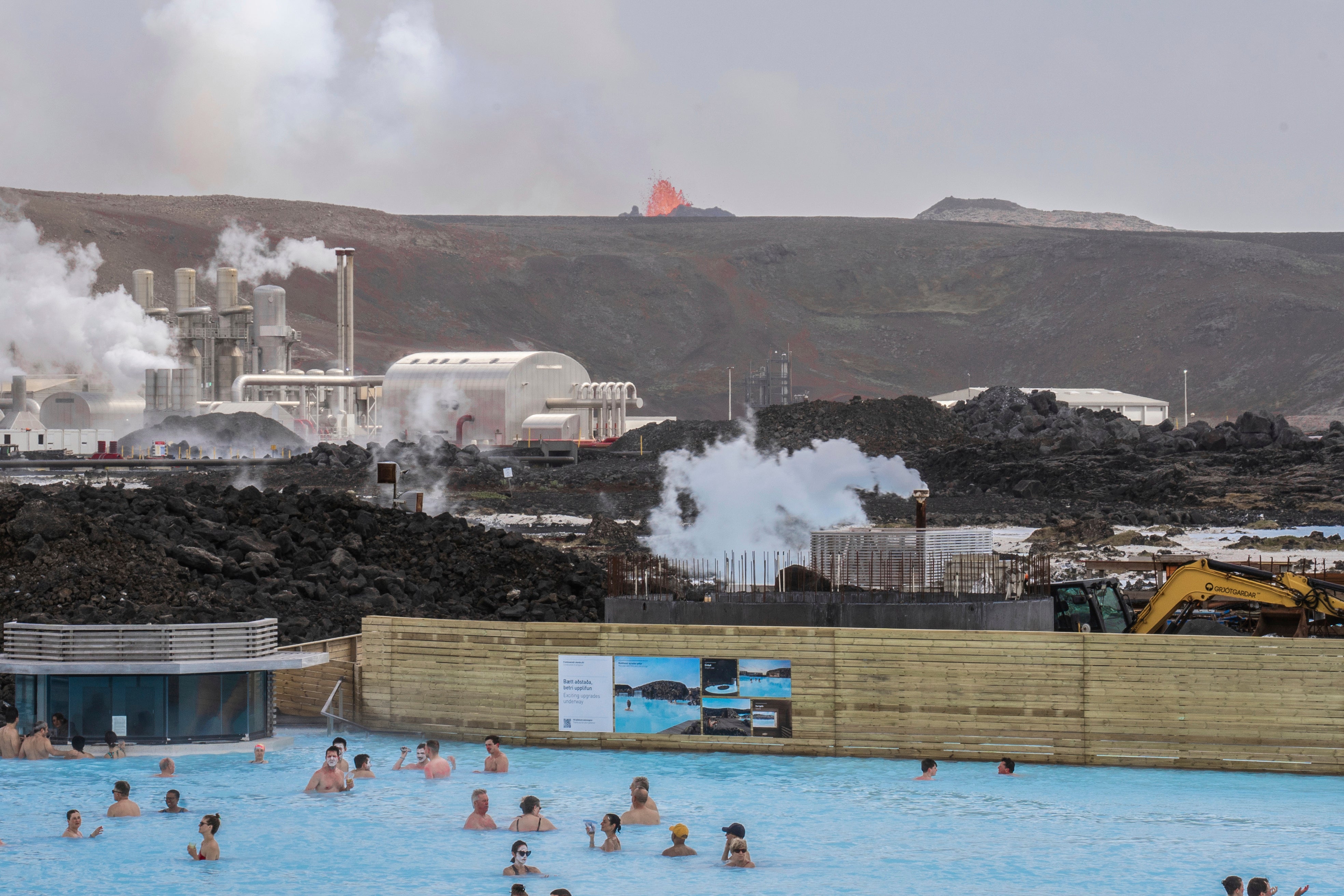 The Blue Lagoon with people bathing in it as the volcanic crater spews lava in the background in Grindavik