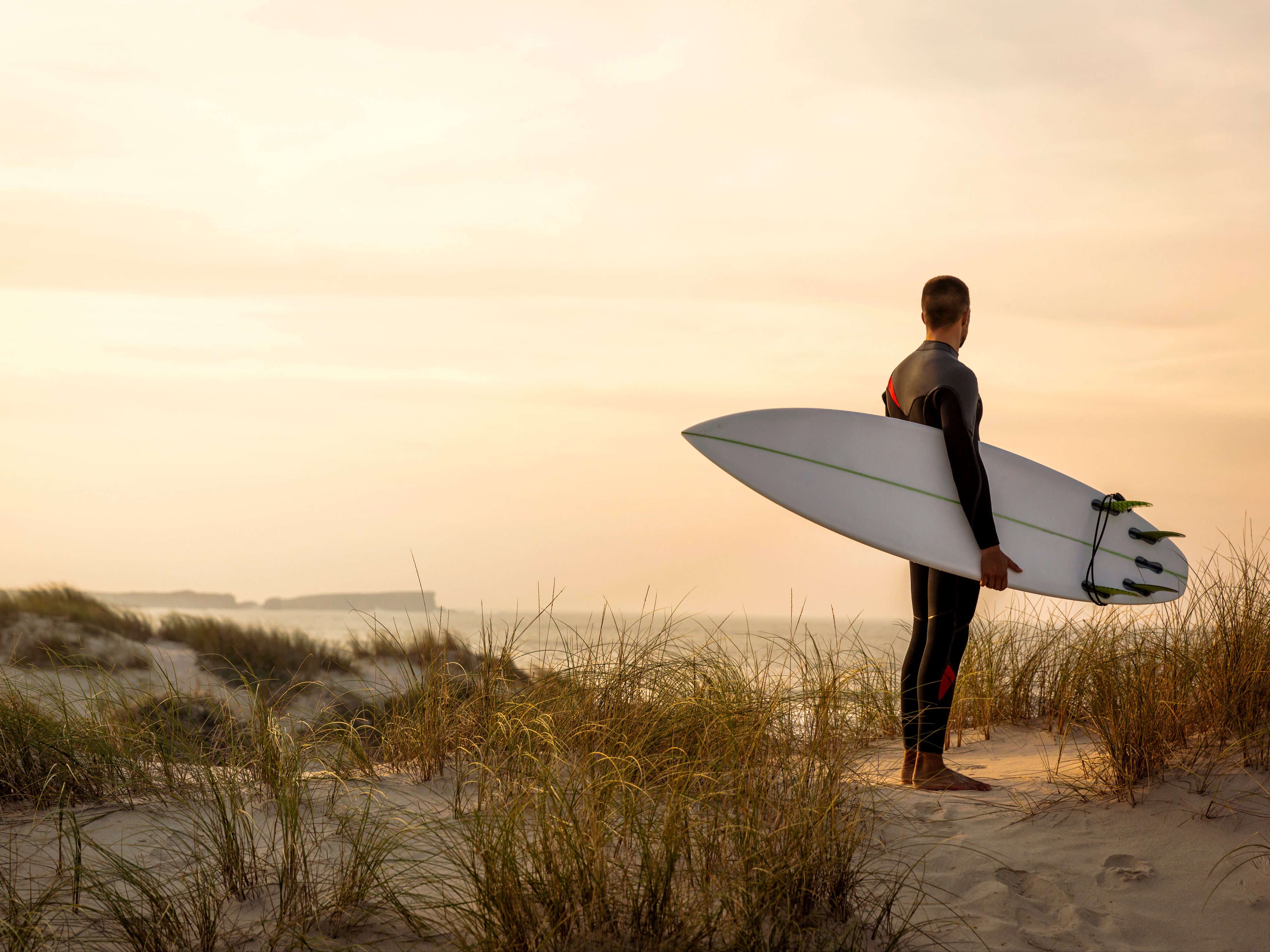 A surfer with his surfboard at the dunes looking to the waves