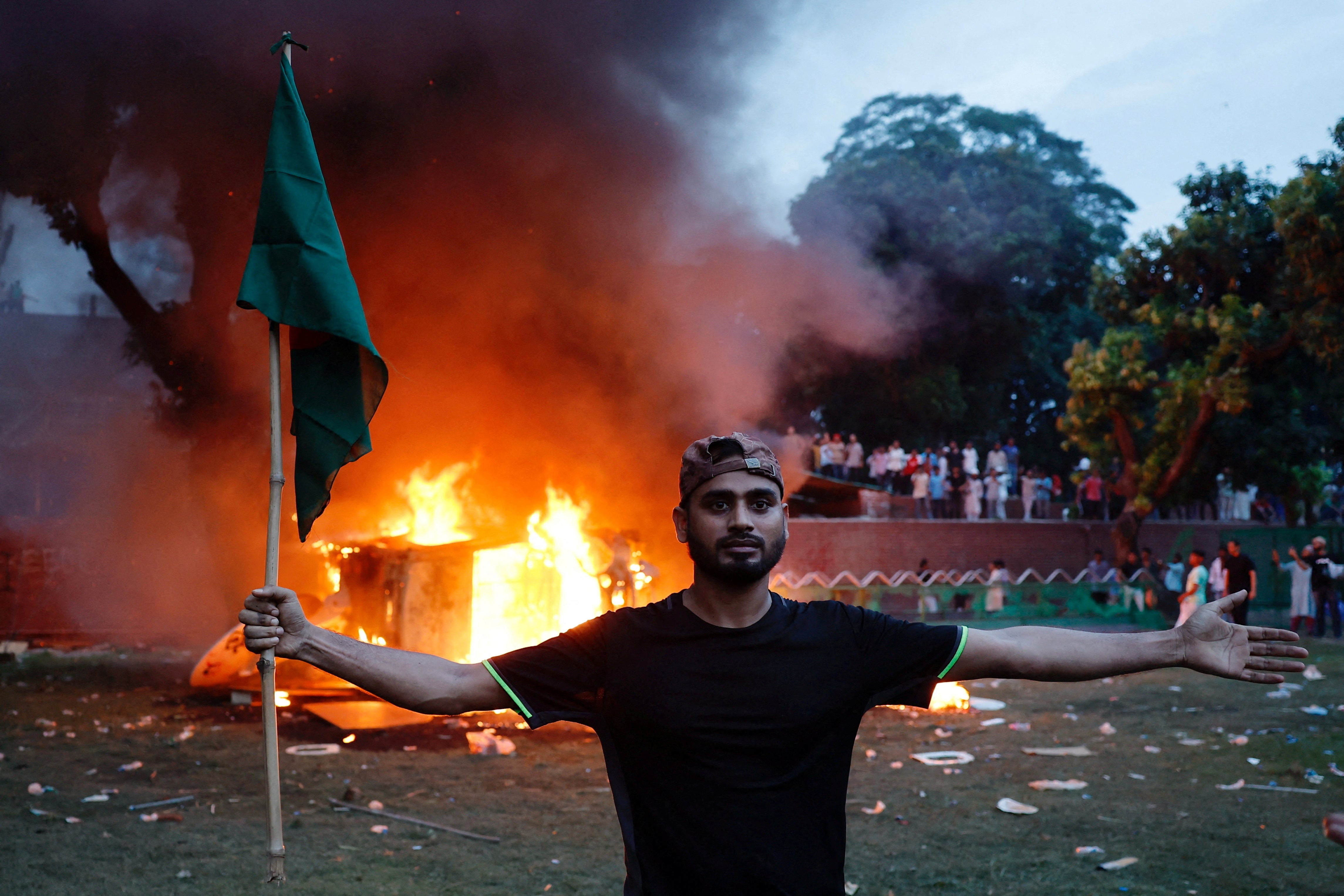 A man holding a Bangladesh flag stands in front of a vehicle that was set on fire at the Ganabhaban, the prime minister’s residence, after the resignation of PM Sheikh Hasina in Dhaka