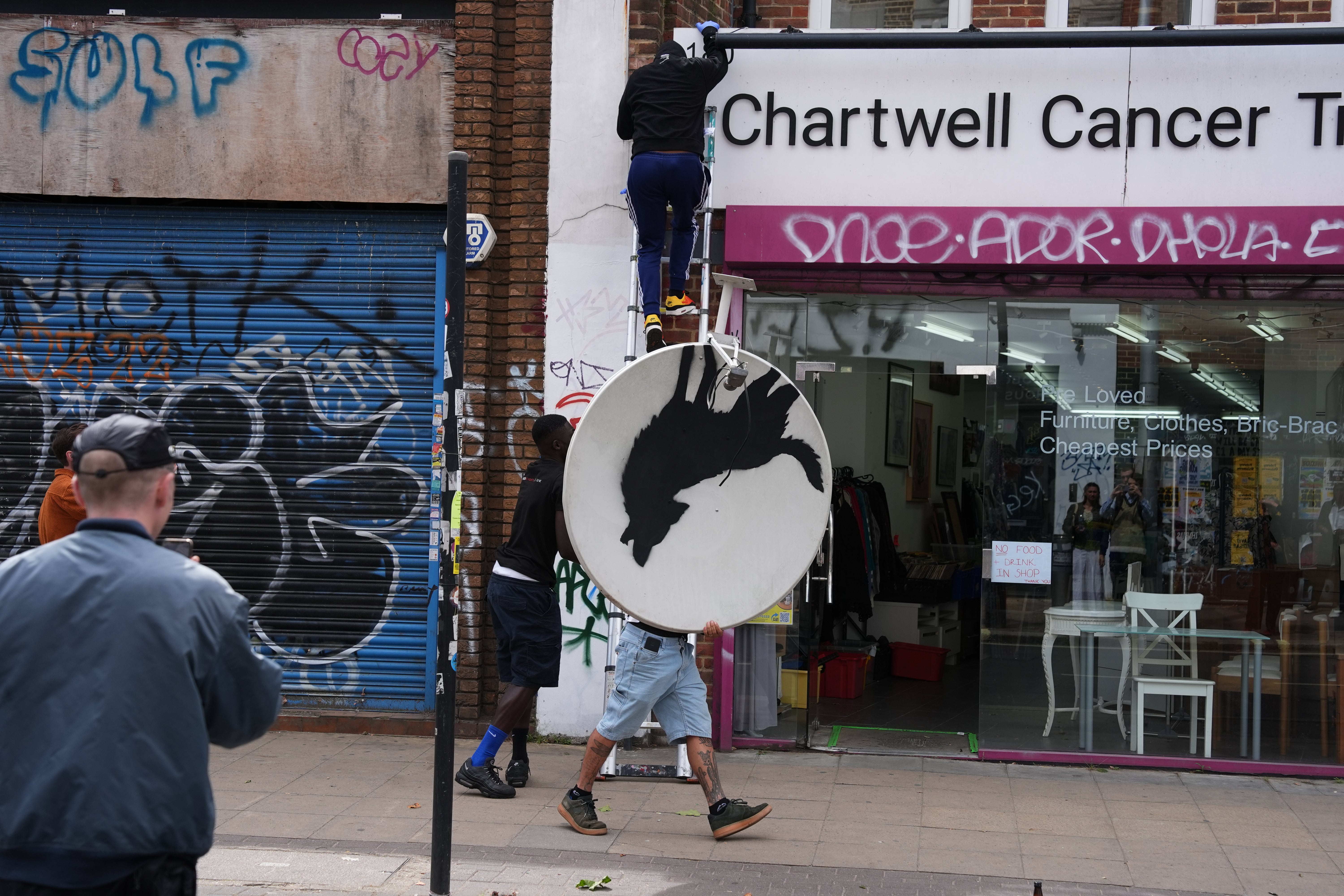 People remove a new artwork by Banksy, depicting a howling wolf painted on a satellite dish that was placed on a shop roof in Peckham, south London