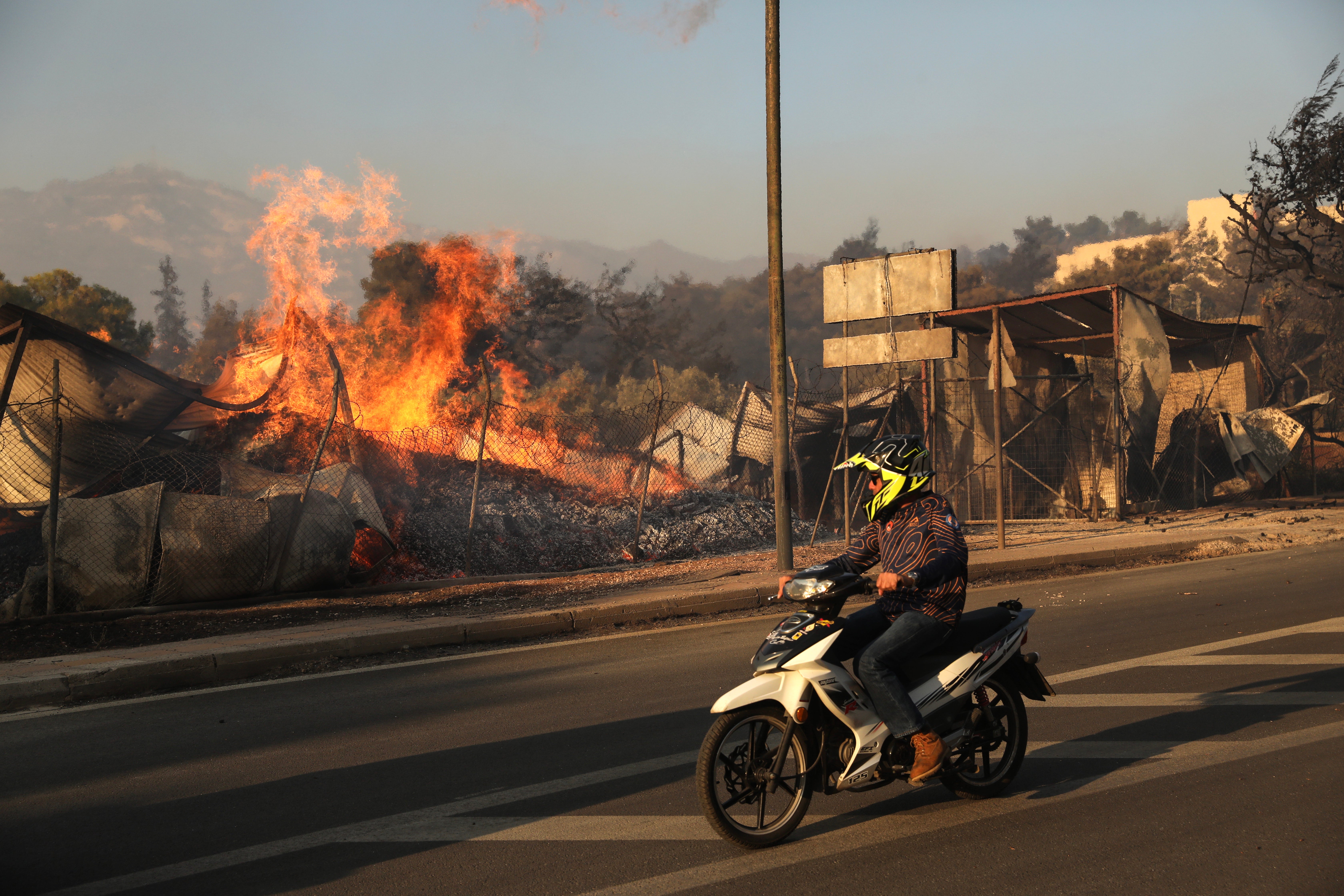 A person riding a scooter watches as a wildfire engulfs a firewood business, in Penteli