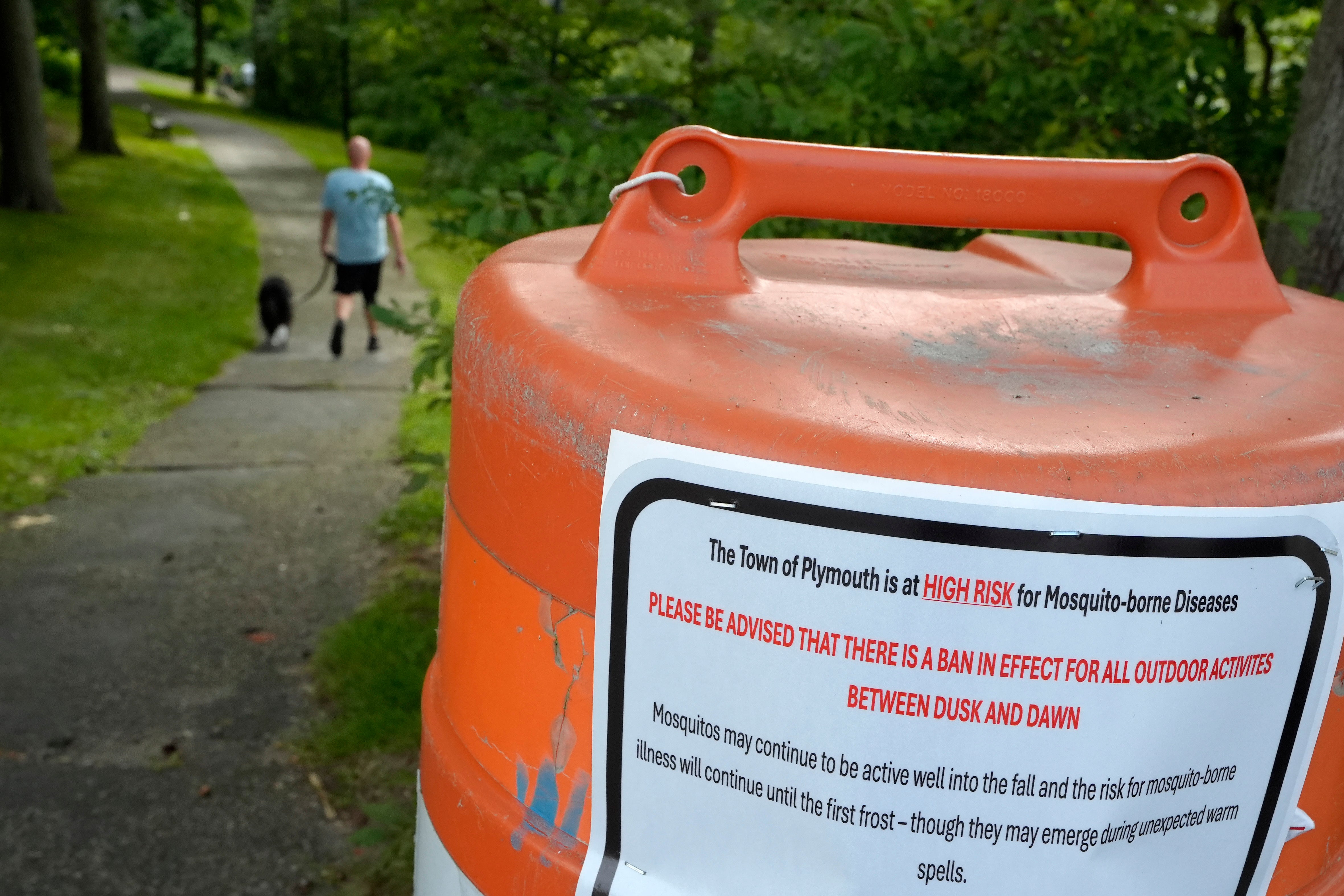 A man walks a dog along a path in Plymouth, Massachusetts. On the path, a sign is posted warning of a curfew related to high risk of mosquito-borne disease. Plymouth is one of the state’s towns that has been sprayed with insecticide, in an effort to stop potential viral infections.