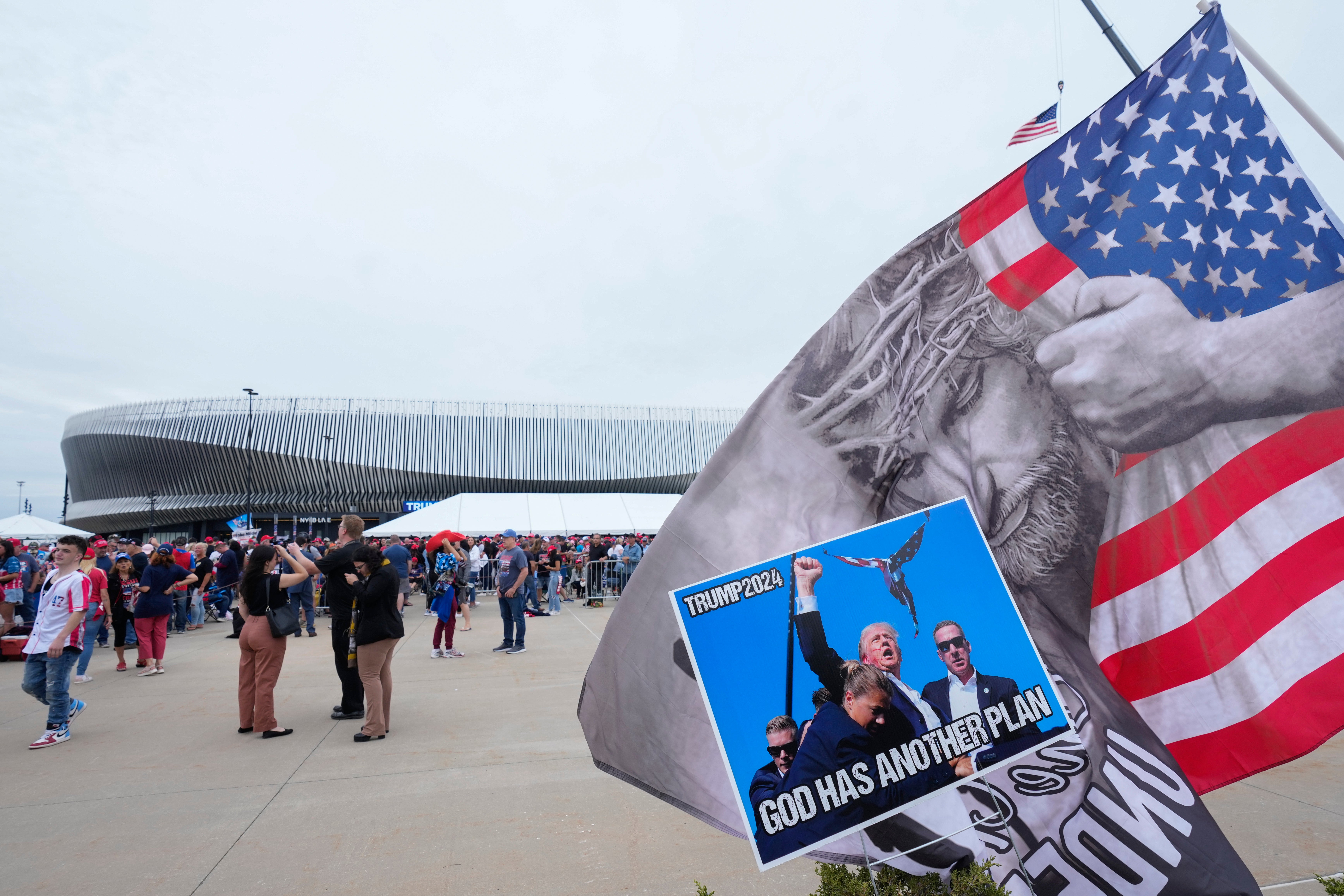 A large flag outside Donald Trump’s rally in Uniondale, New York, shows what appears to be Jesus Christ hugging or peeling back an American flag. Next to the flag is a version of the iconic image of Trump after his first assassination attempt with what appears to be some kind of winged creature — perhaps an angel — hovering in the air above him, with the words “God Has Another Plan” across the bottom