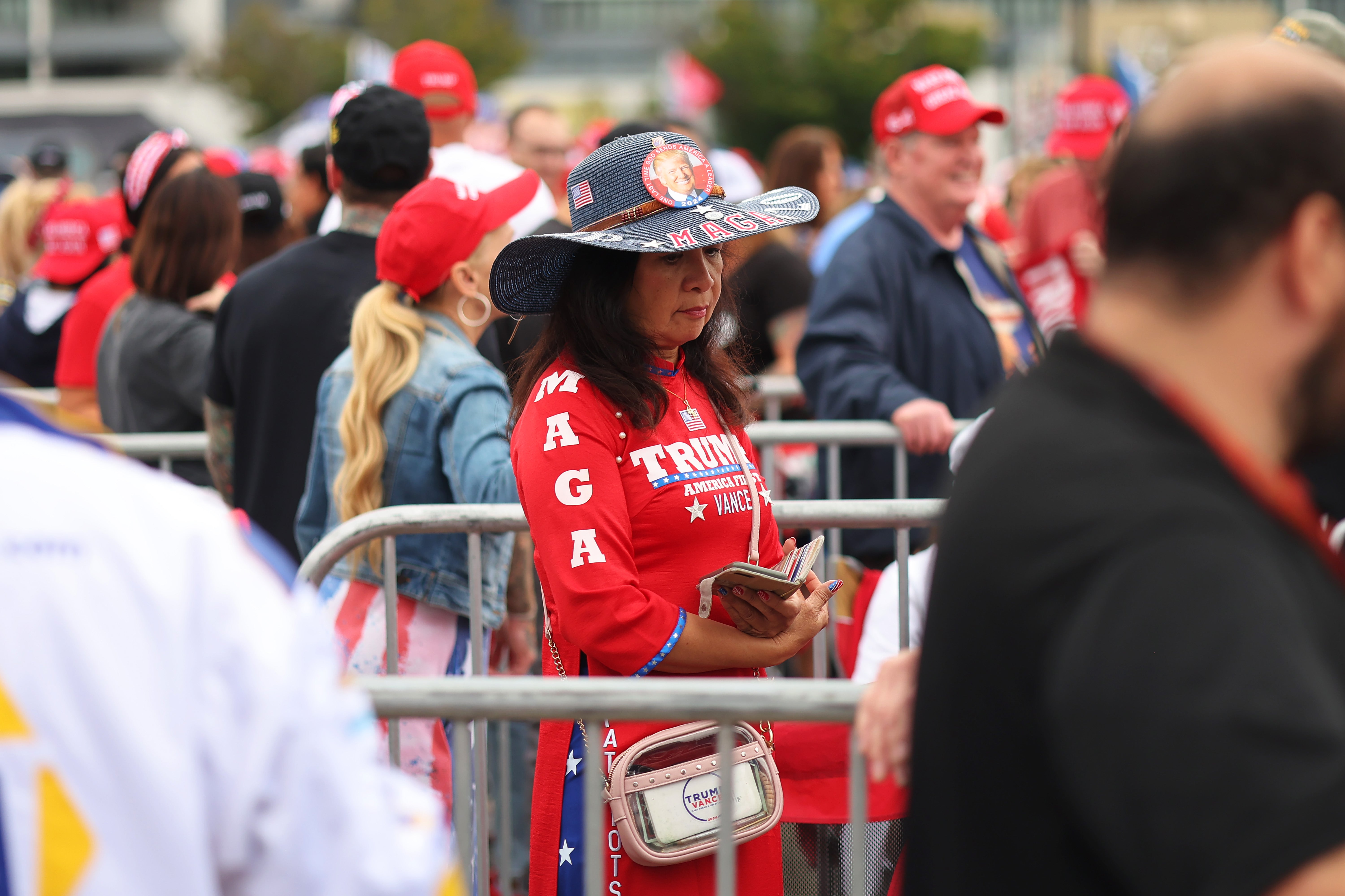 A woman is fully decked-out in Trump gear while waiting to attend his rally. She’s sporting a Trump shirt, pants, purse, floppy cowboy hat and a big button of his grinning face on the hat’s crown