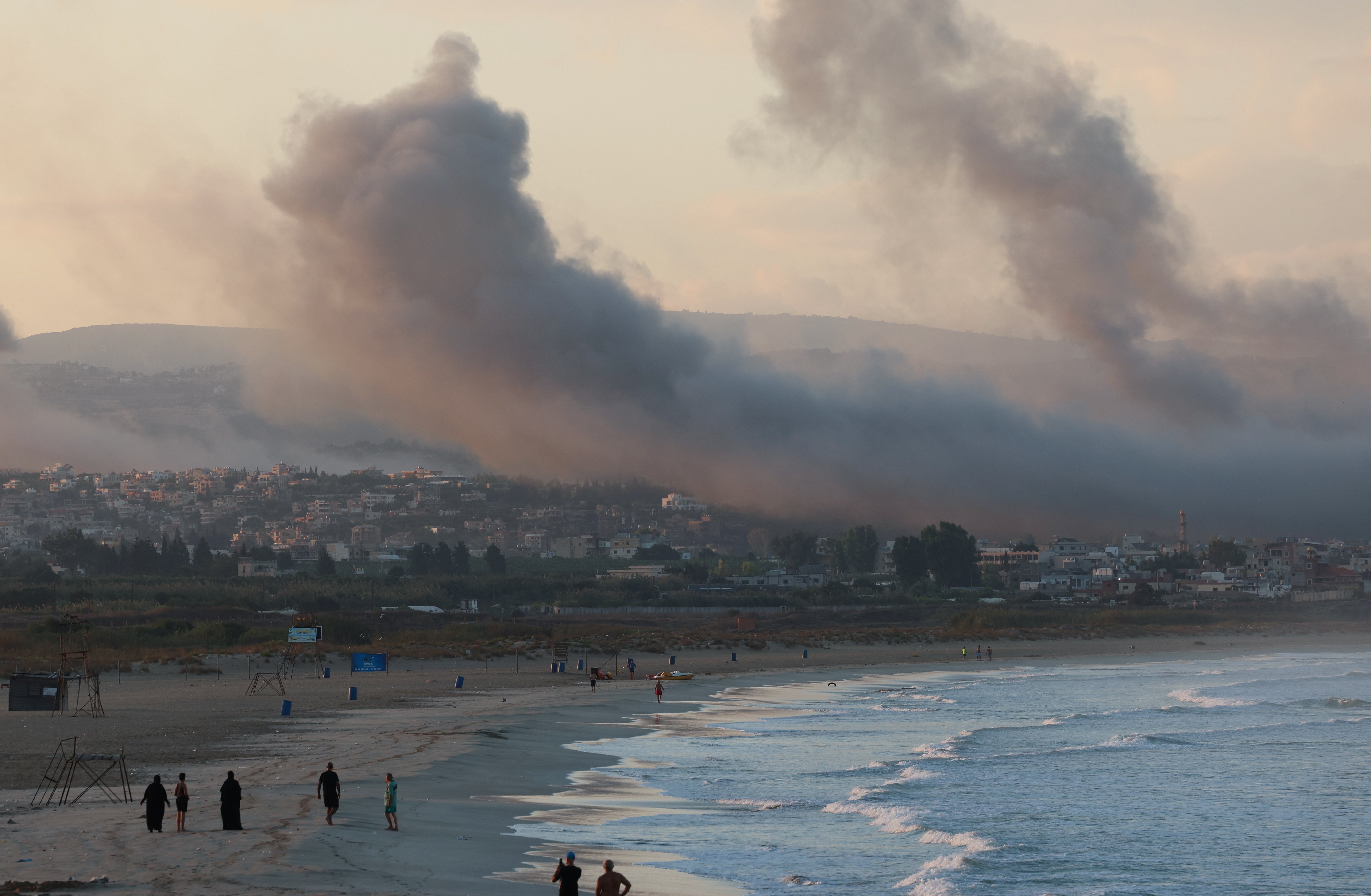 People walk at a beach as smoke billows over southern Lebanon following Israeli strikes