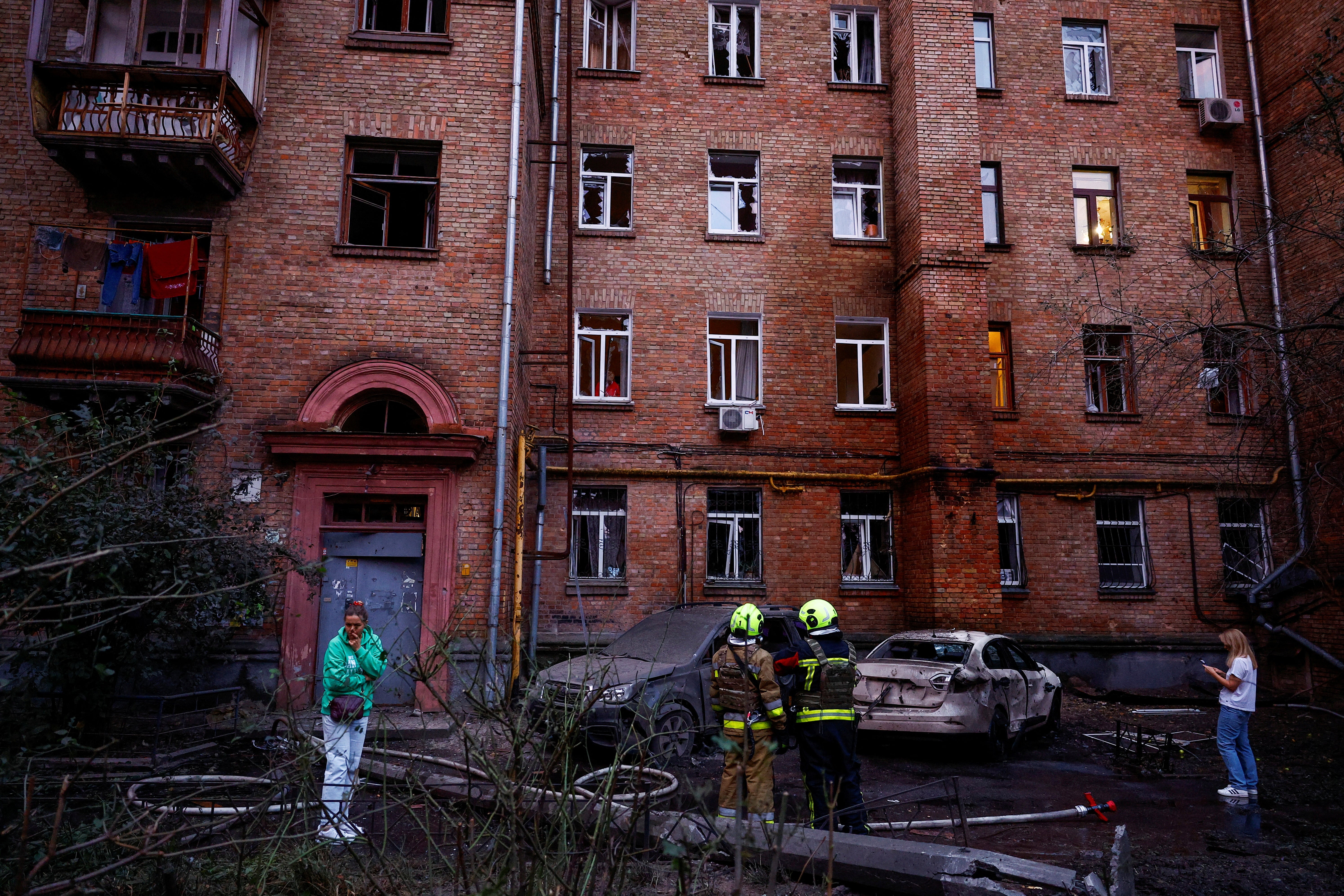 Residents and firefighters stand next to cars and an apartment building damaged during a Russian drone strike in Kyiv