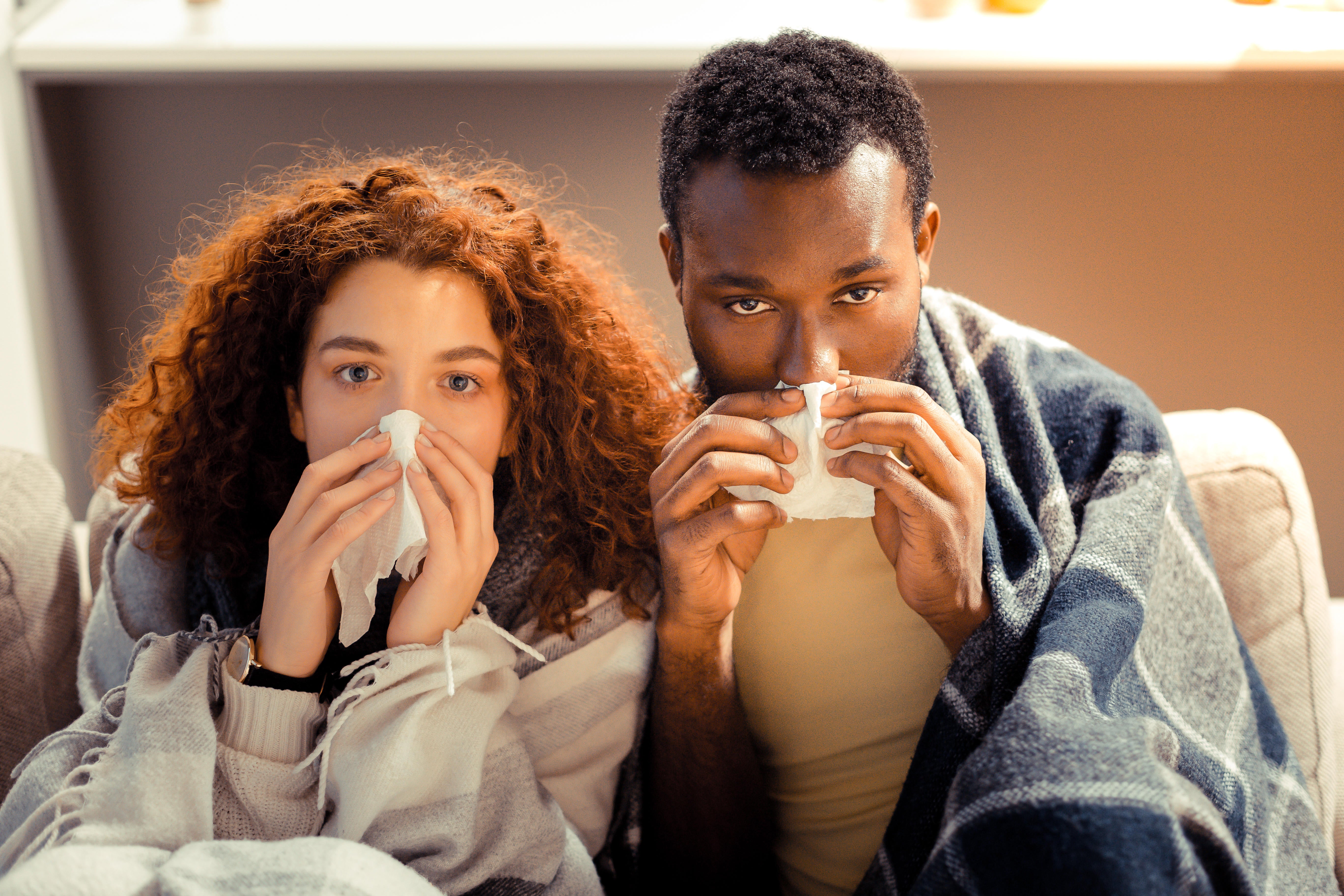 A man and woman sat on the sofa at home, wrapped in blankets and blowing their noses into a tissue