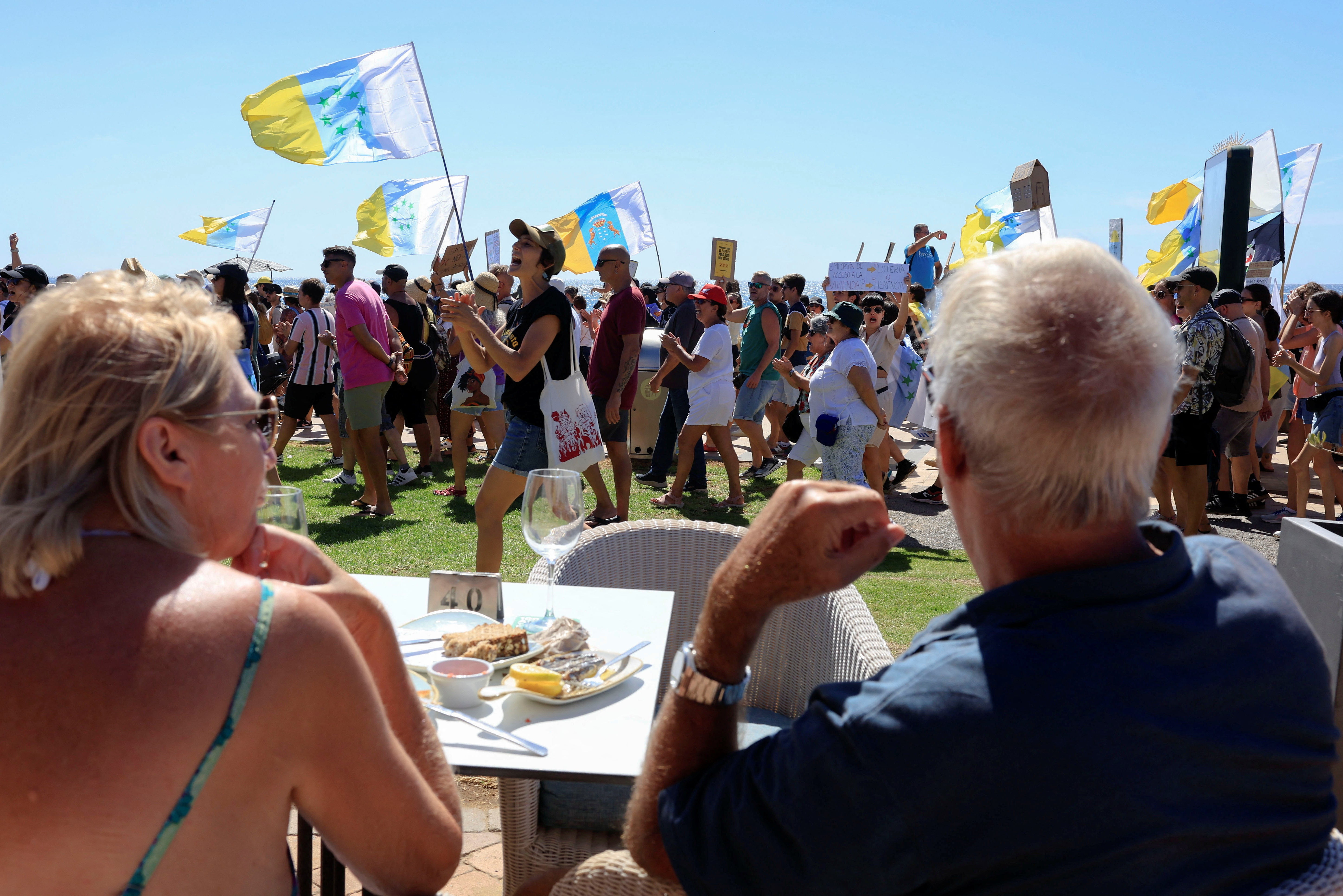 A couple of tourists sitting on a terrace watch a demonstration for a change in the tourism model in the Canary Islands