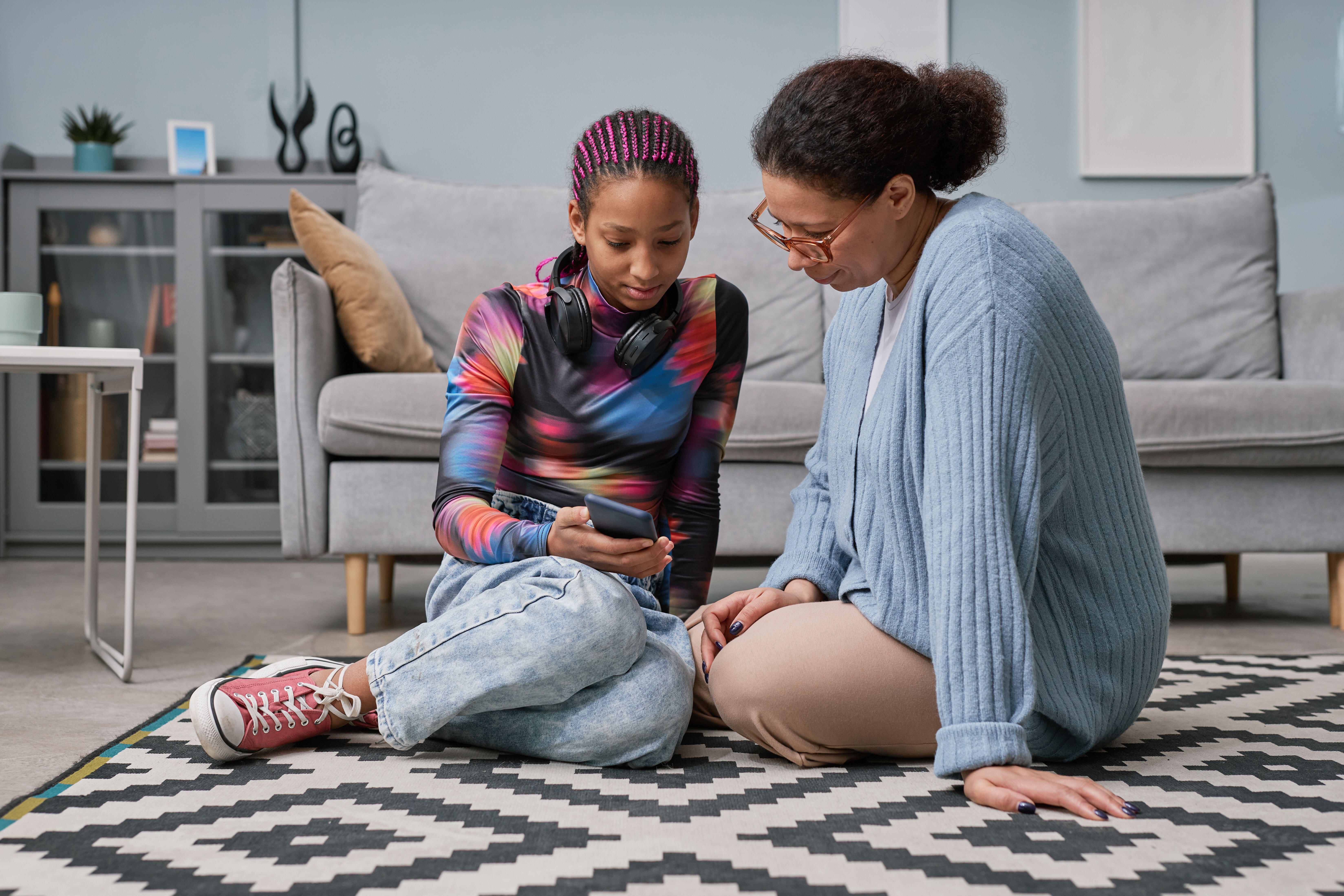 Teenage girl sat on the floor with her mum looking through her phone (Alamy/PA)