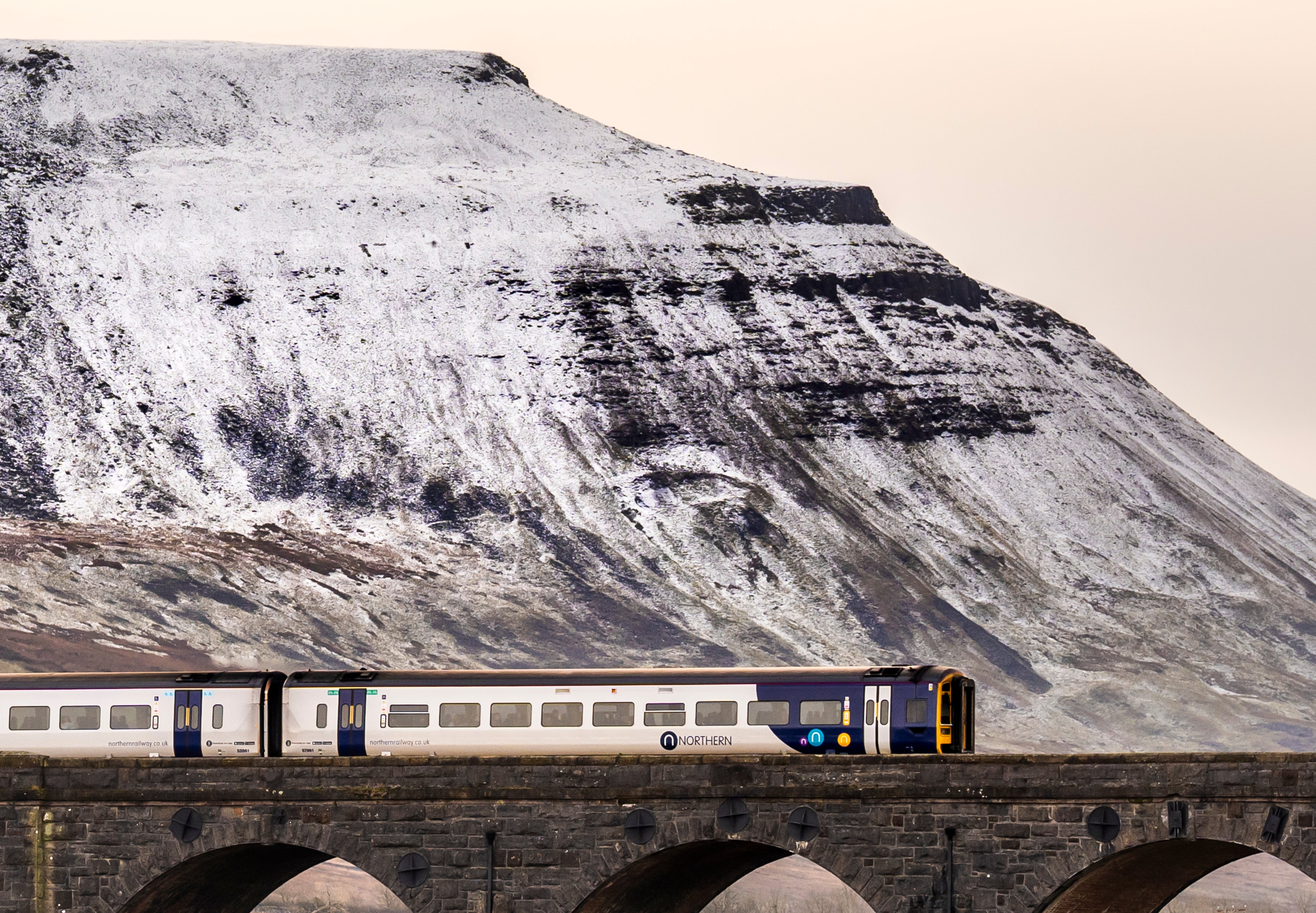 A train crosses over the Ribblehead Viaduct with the snow capped mountain of Ingleborough behind, in the Yorkshire Dales.